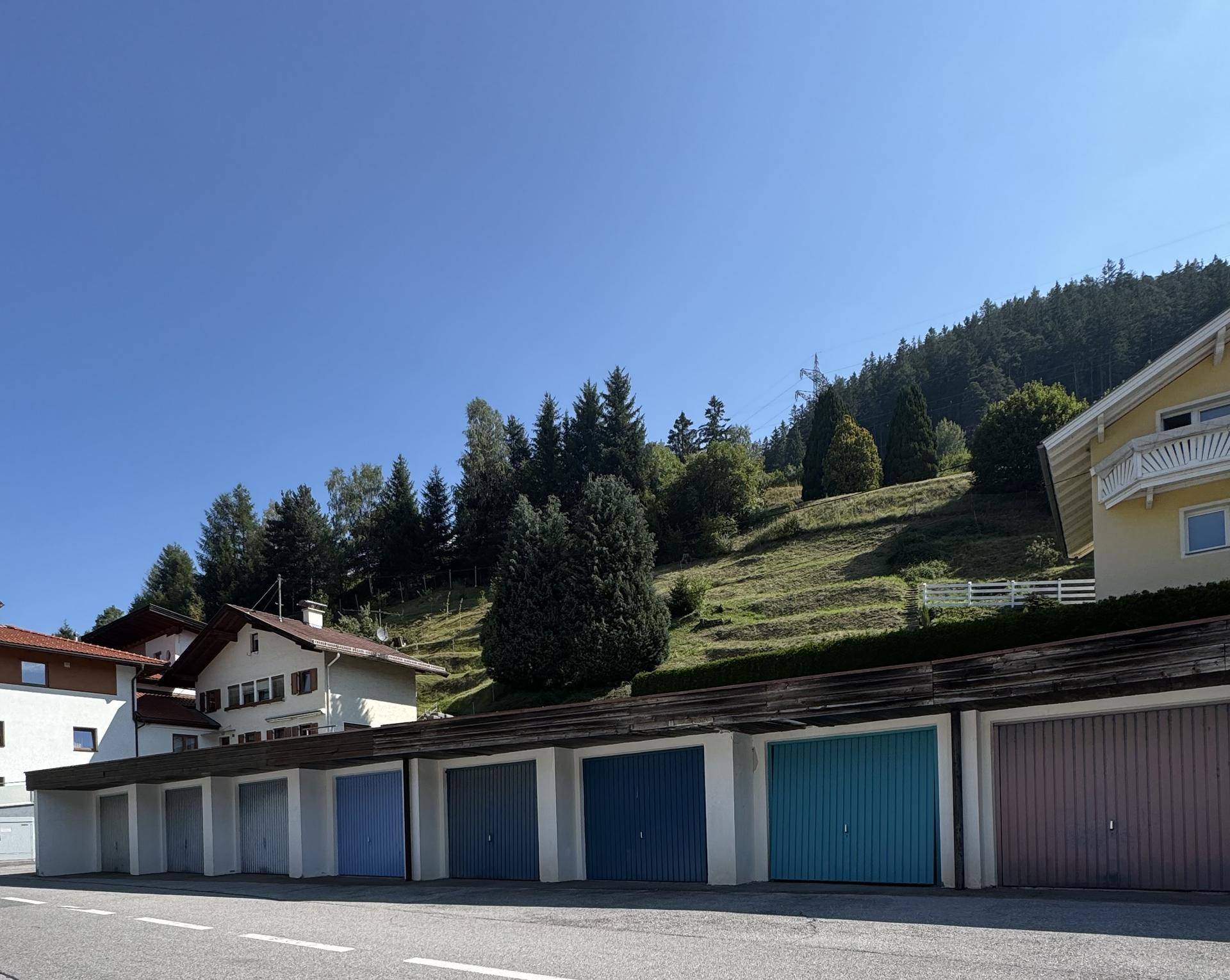 Reihe von Garagen mit bunten Toren vor einem grünen Hügel mit Bäumen und Häusern.