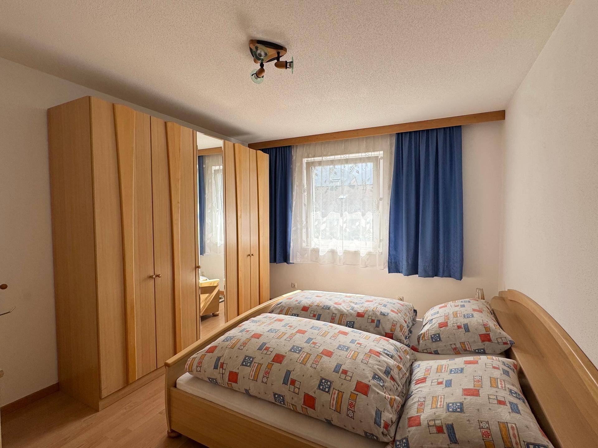 Gemütliches Schlafzimmer mit Holzschrank, Doppelbett und Fenster mit blauen Vorhängen.