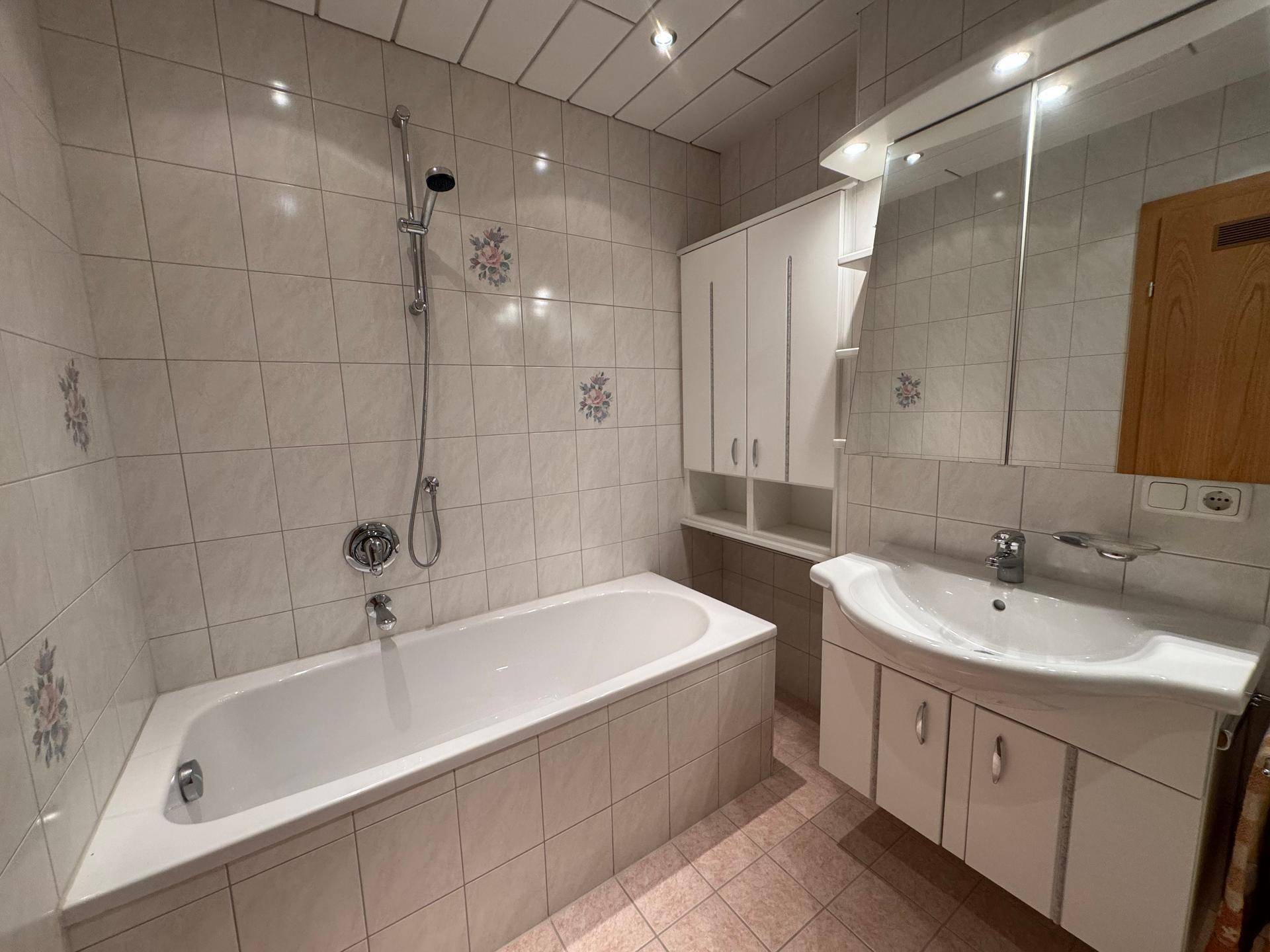 Helles Badezimmer mit Badewanne, Waschbecken und Spiegelschrank, gefliest in hellen Tönen.