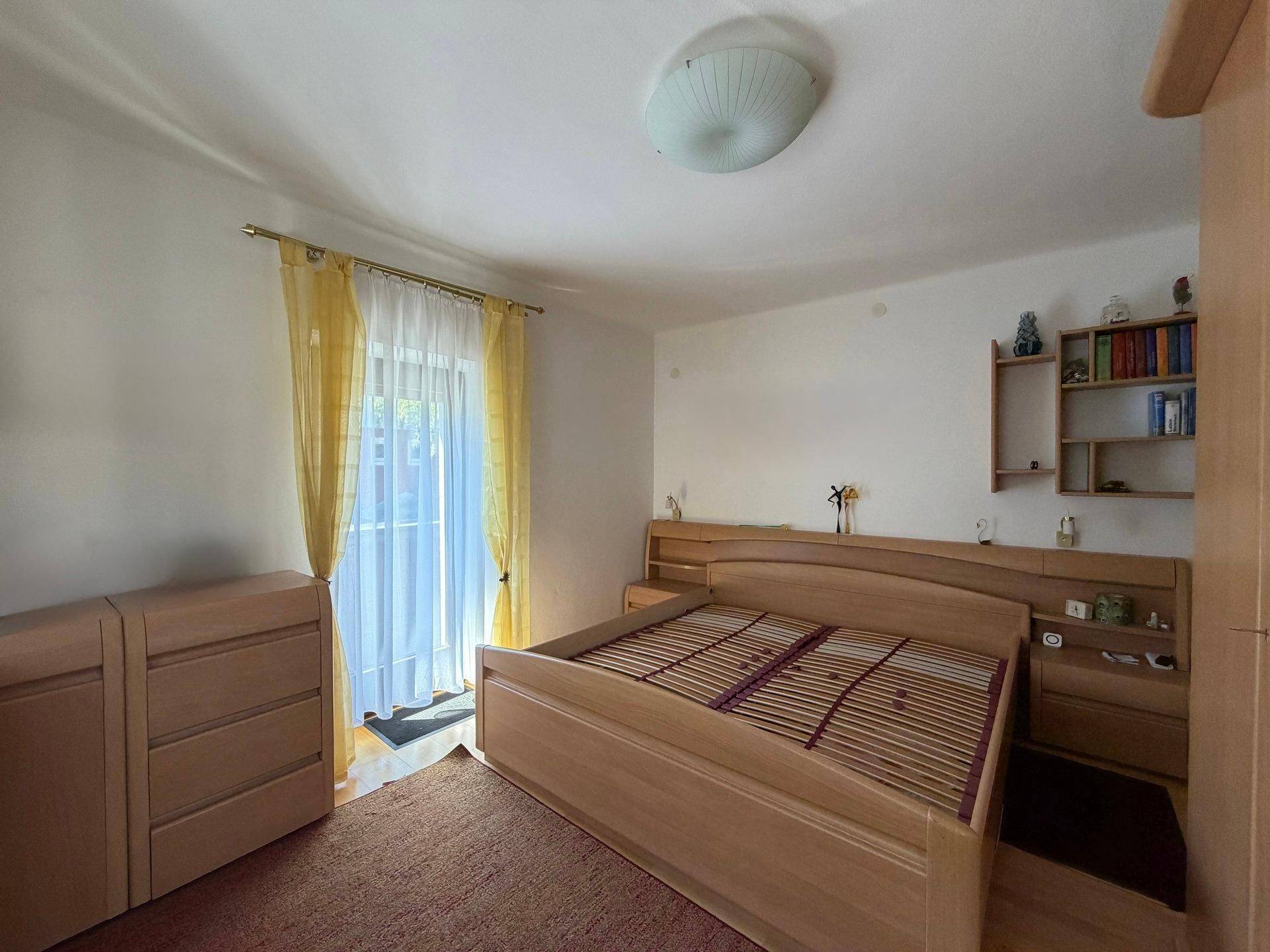 Schlafzimmer mit Doppelbett, Holzmöbeln und Zugang zu einem Balkon mit viel Tageslicht.