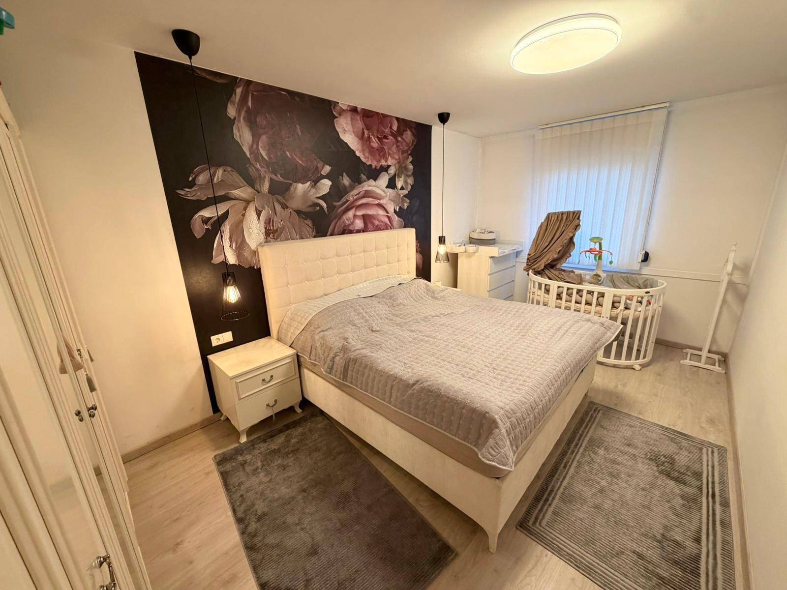 Gemütliches Schlafzimmer mit floraler Akzentwand, großem Bett und Babybettchen.