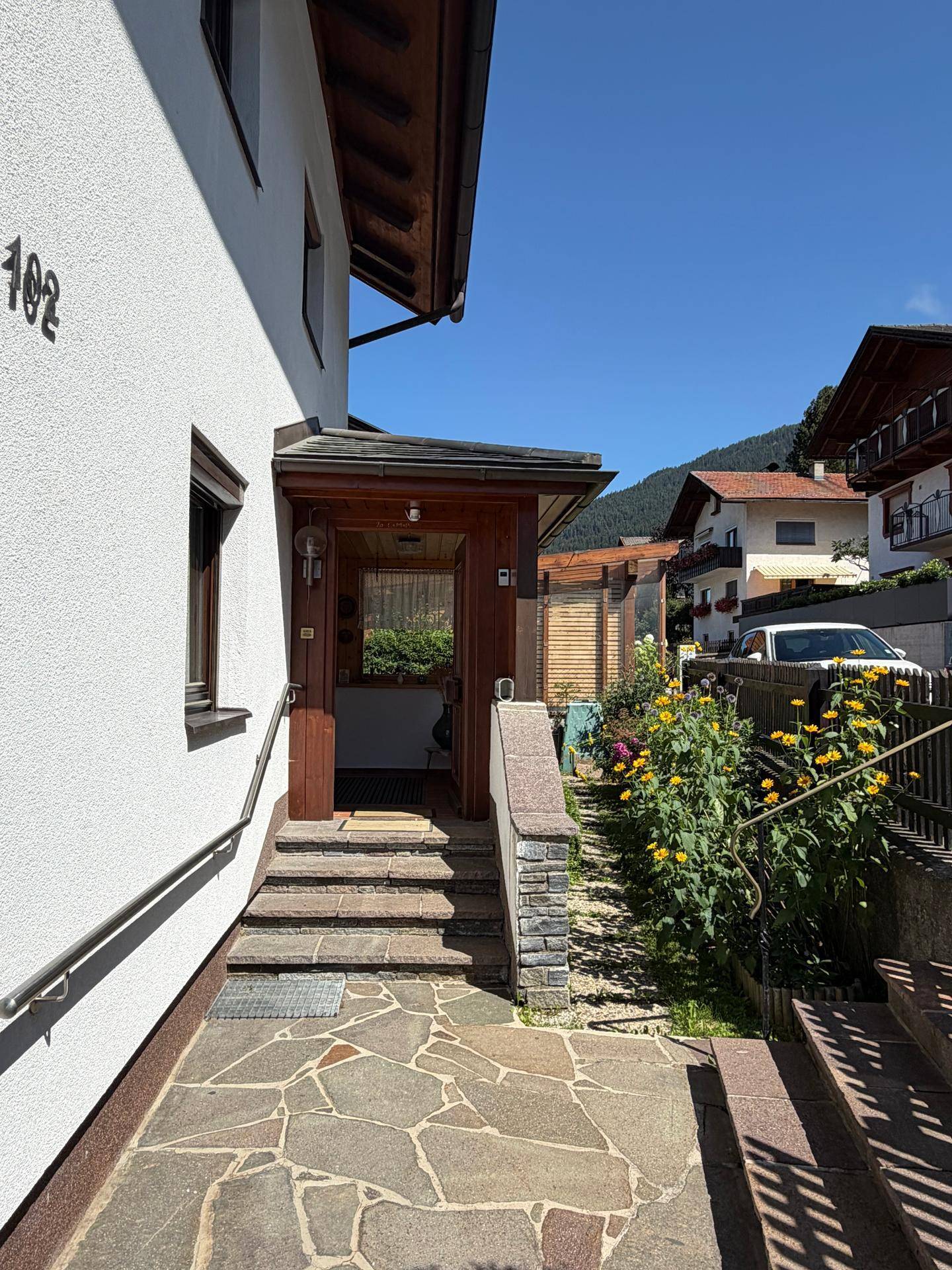 Eingangsbereich des Hauses mit Steintreppe, Handlauf und blühenden Pflanzen im Vorgarten.
