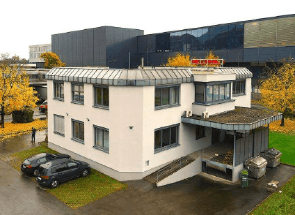 Seitenansicht des Bürogebäudes mit Laderampe und Müllcontainern, umgeben von Herbstbäumen.