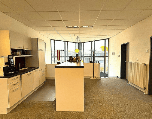 Helle Büroküche mit Küchenzeile und Theke, großen Fenstern und Teppichboden, ideal für Pausen.