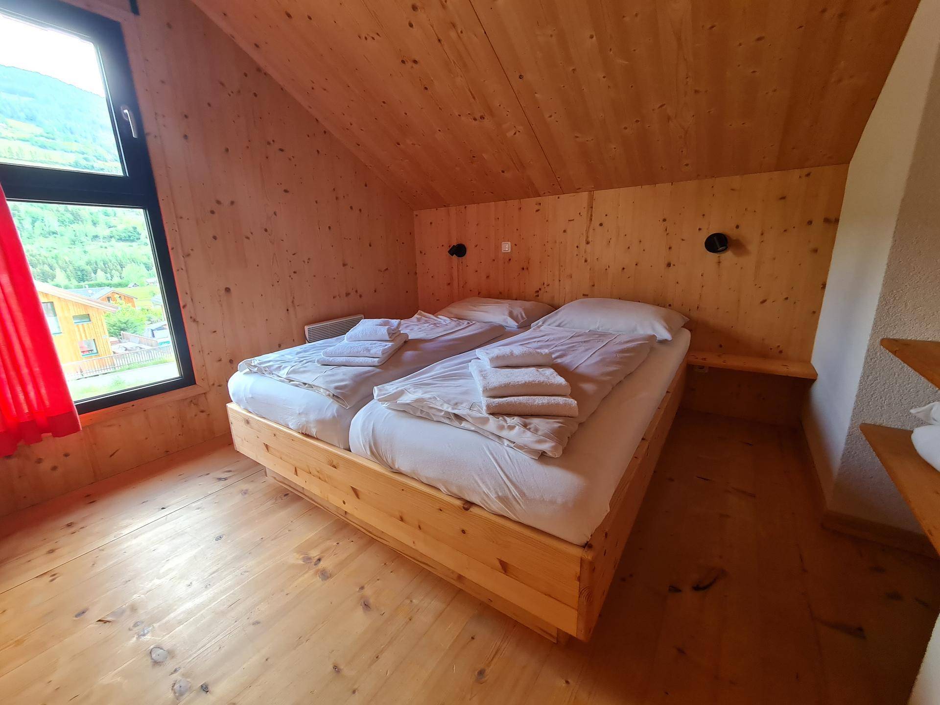 Schlafzimmer im Dachgeschoss mit Holzwänden, Doppelbett und Fenster mit rotem Vorhang.