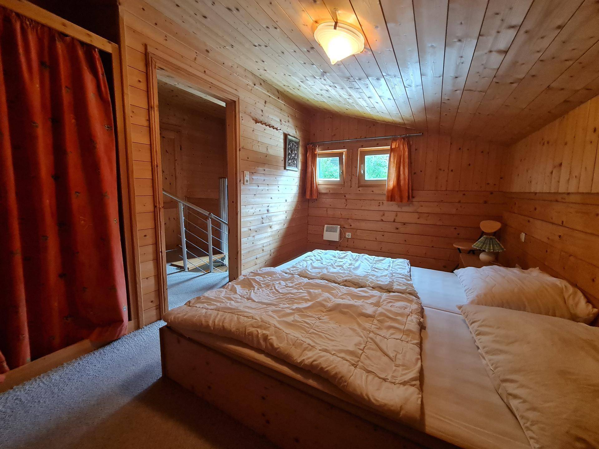 Helles Schlafzimmer mit Doppelbett, Holzwänden und zwei kleinen Fenstern, mit Zugang zum Flur.