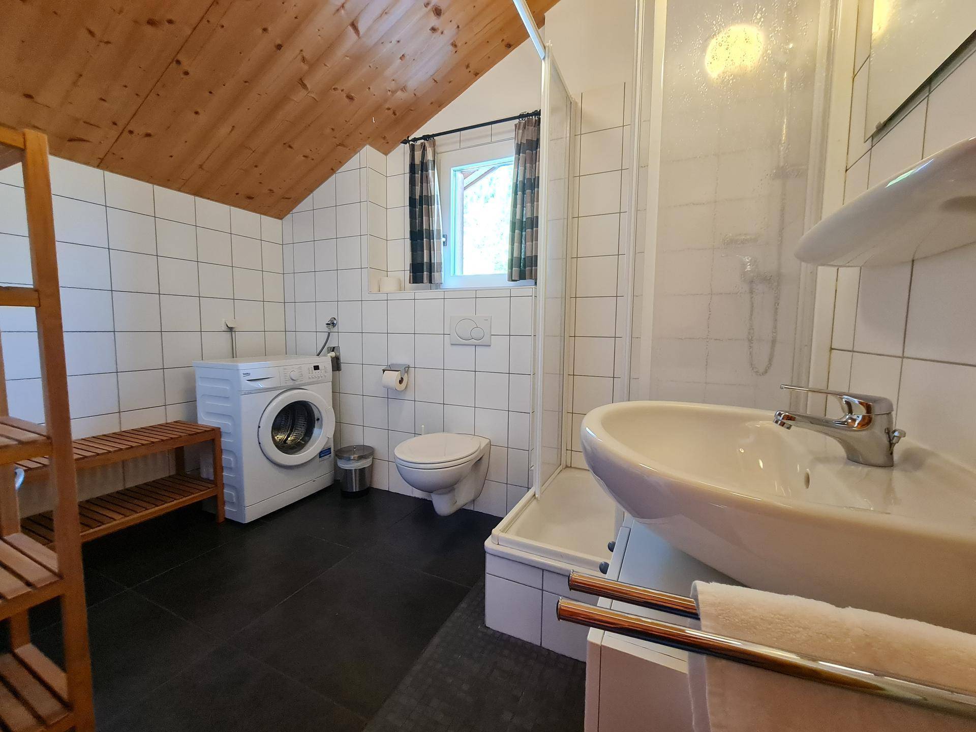 Badezimmer mit Dusche, Waschmaschine, WC und Fenster im Dachgeschoss.