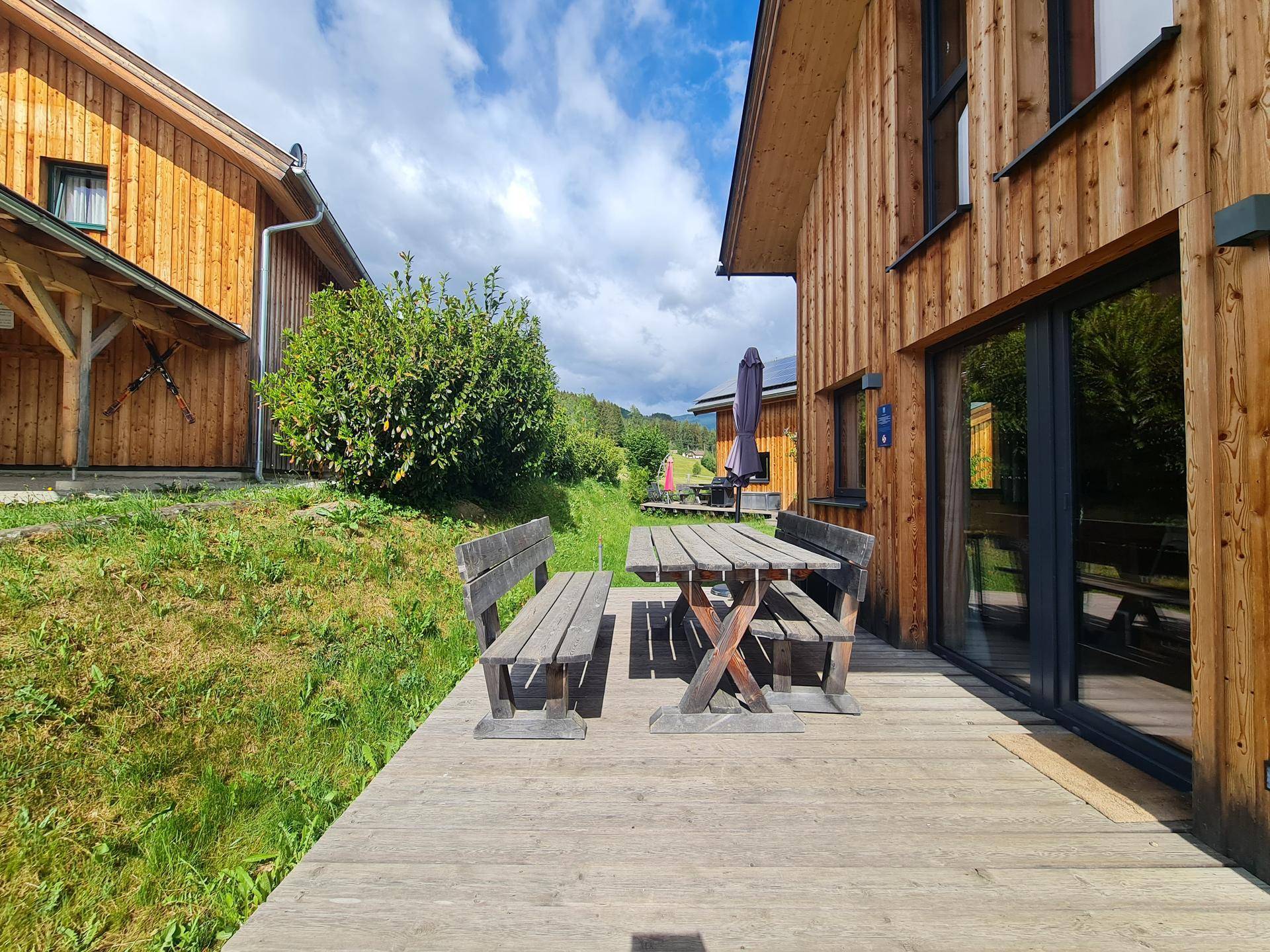 Holzterrasse mit rustikalem Tisch und Bänken, umgeben von grüner Natur und Sonnenschein.
