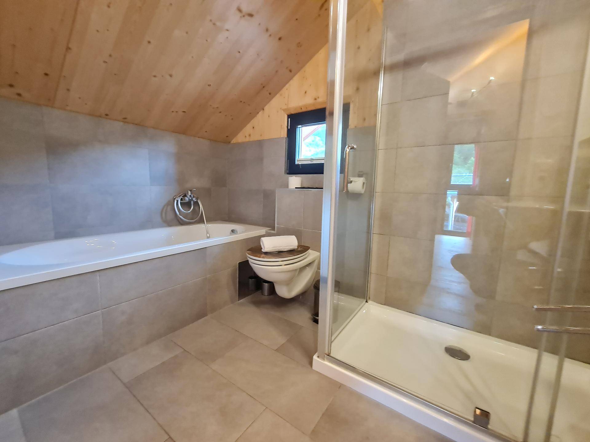 Helles Badezimmer mit Badewanne, Duschkabine und grauen Fliesen im Dachgeschoss.