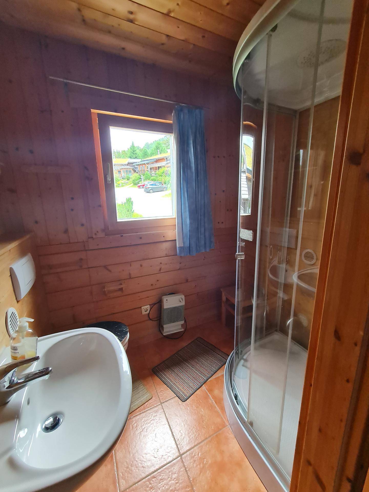 Kleines Badezimmer mit Duschkabine, Waschbecken und Fenster, komplett in Holz verkleidet.