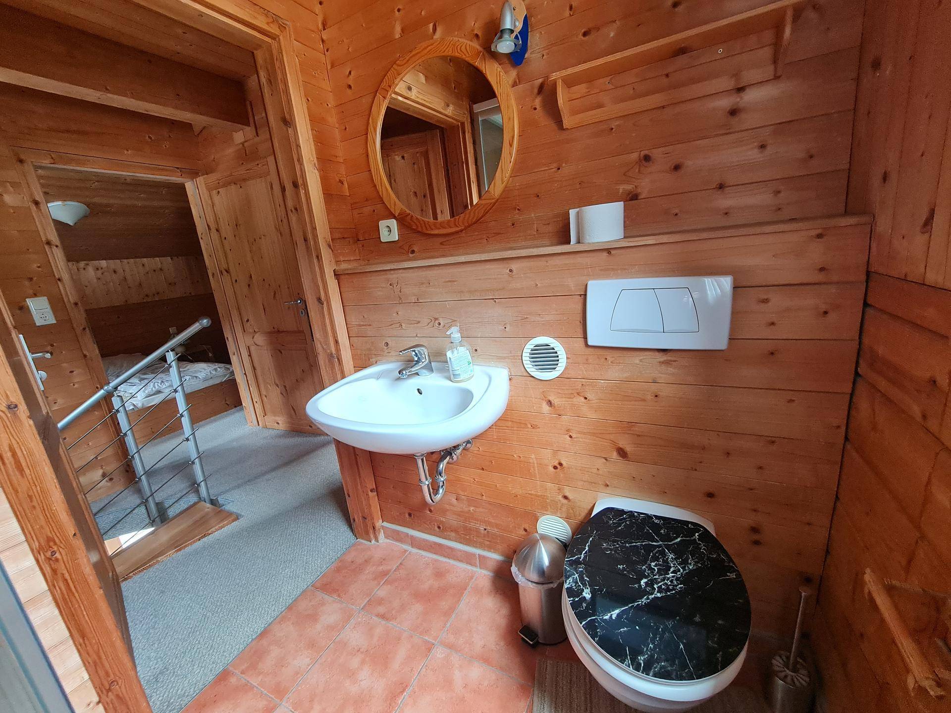 Separates WC mit Waschbecken, Spiegel und Holzwänden, funktional und einfach gehalten.