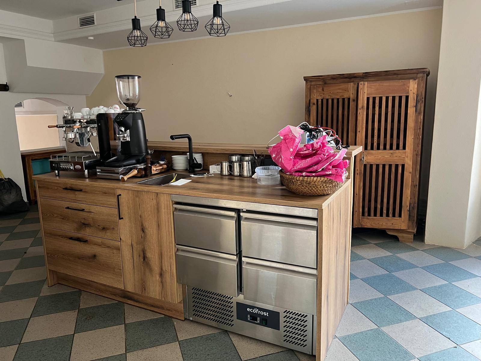 Holztheke mit Kaffeemaschine und Kühlschubladen in einem Gewerberaum mit kariertem Fliesenboden.