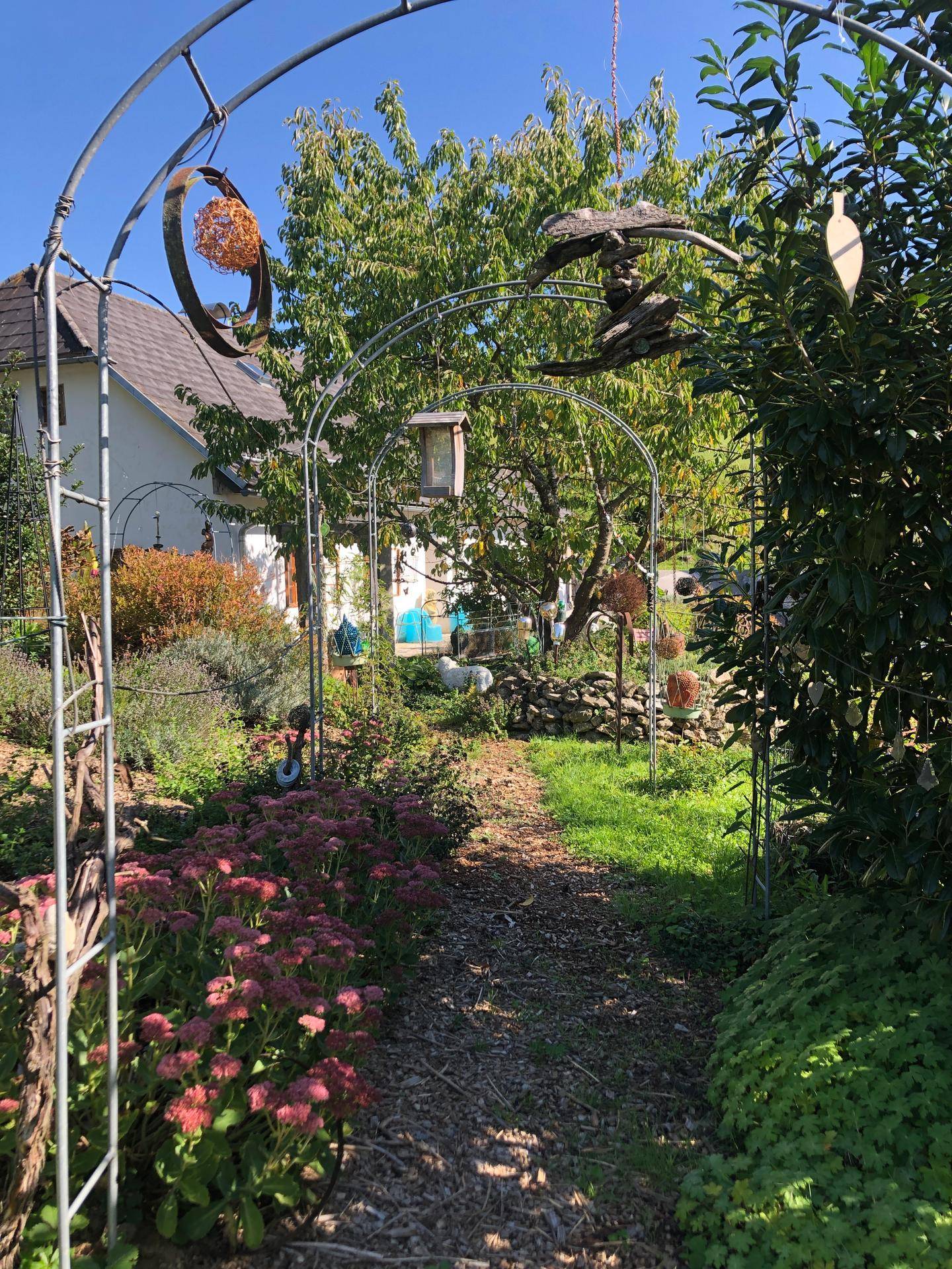 Idyllischer Gartenweg unter einem Metallbogen, umgeben von üppiger Vegetation und Blumen.