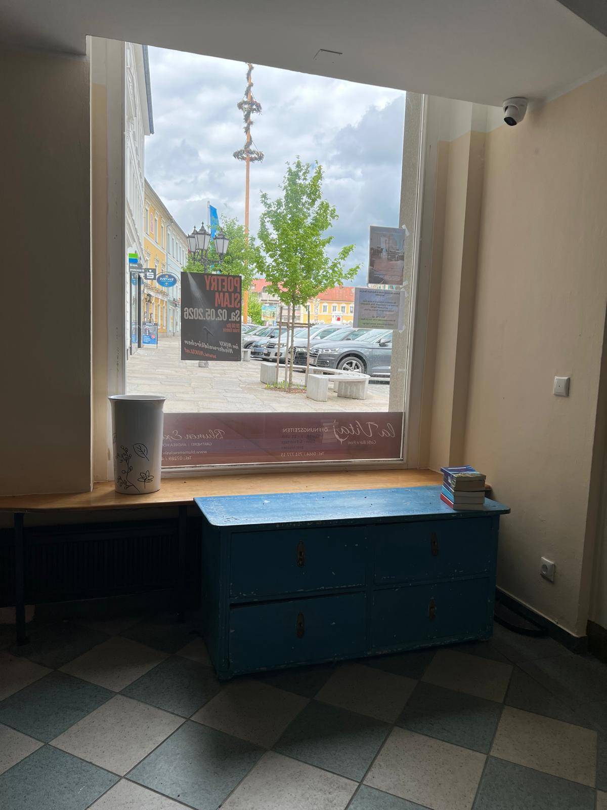 Innenansicht mit Blick durch ein großes Fenster auf eine belebte Straße und einen blauen Holzkasten.