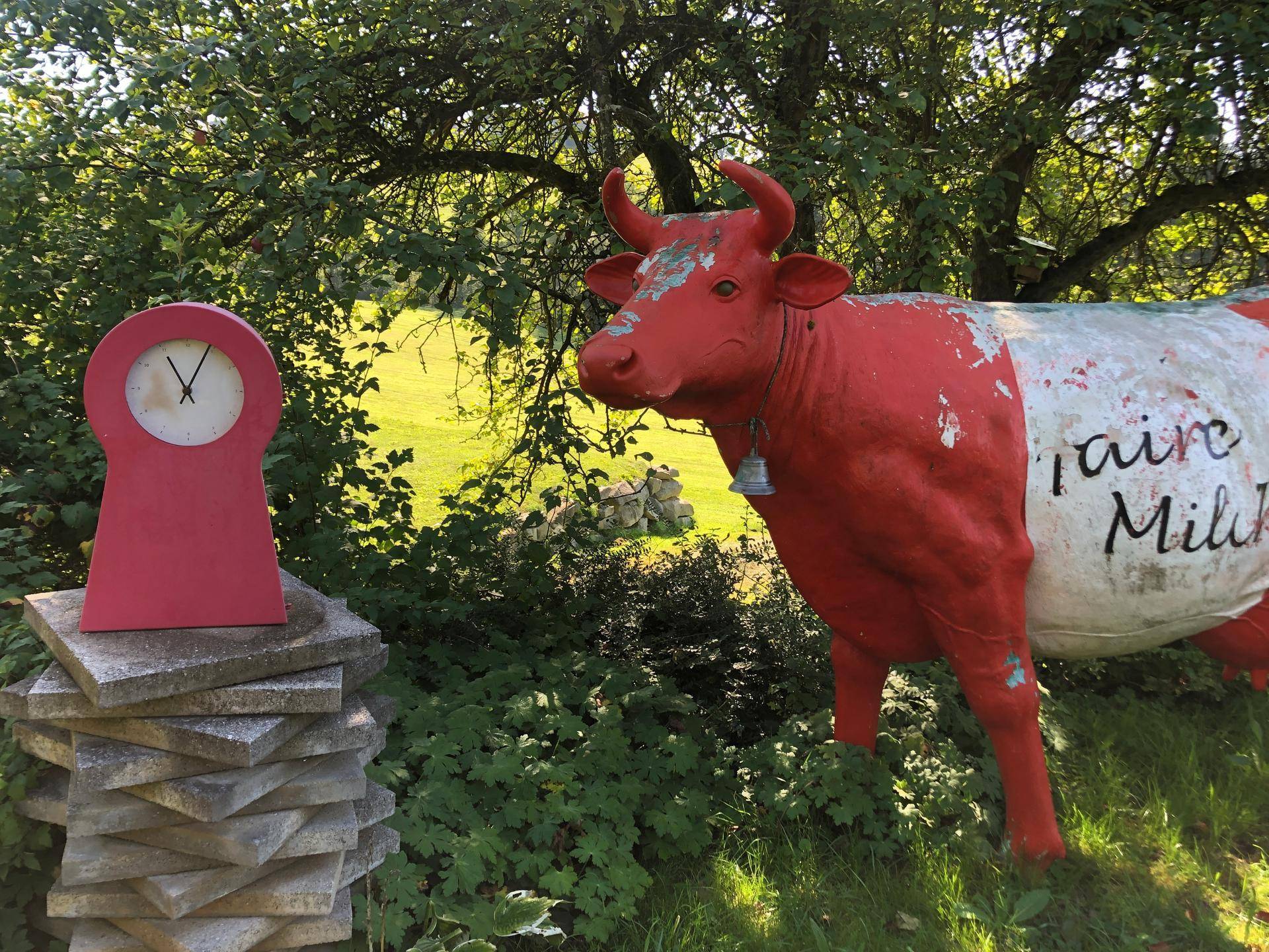 Künstlerische rote Kuhstatue und eine Uhr in einem weitläufigen, grünen Garten.