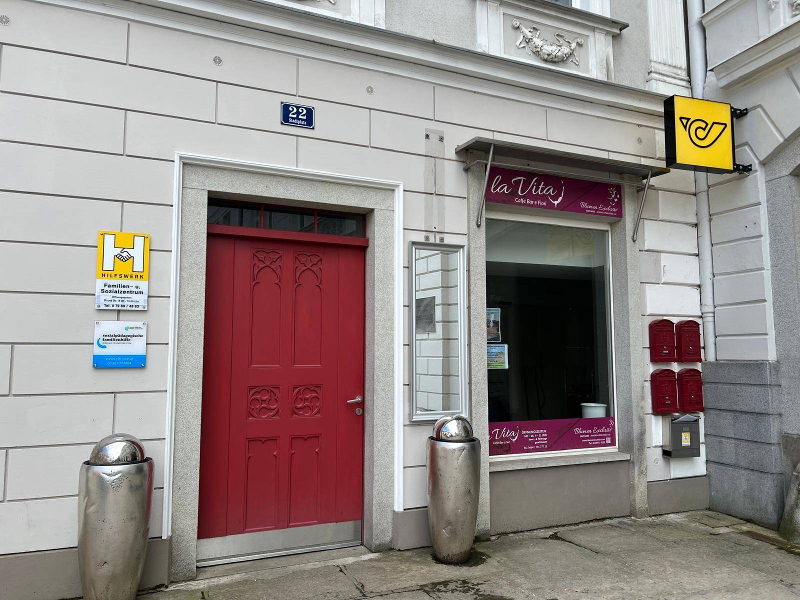Fassade eines Gebäudes mit roter Eingangstür, Schaufenster und verschiedenen Geschäftsschildern.