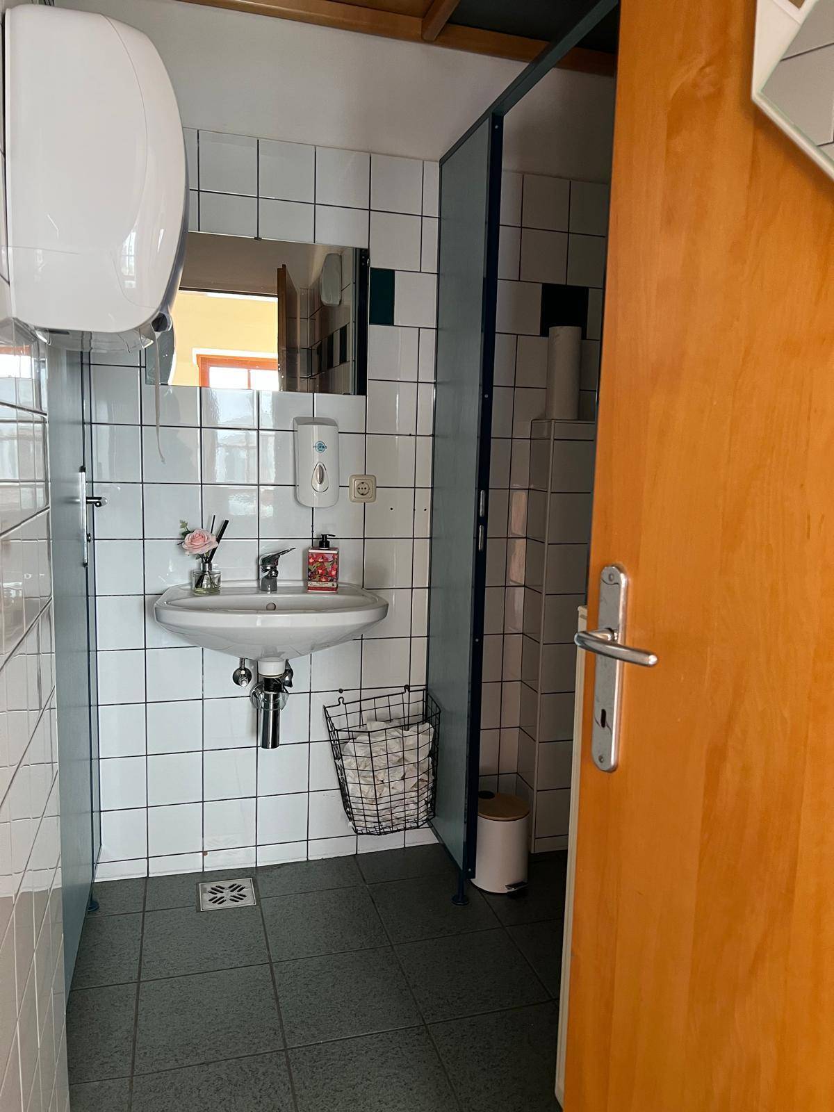 Öffentliche Toilette mit weißen Fliesenwänden, Waschbecken und einer Trennwand für eine Kabine.