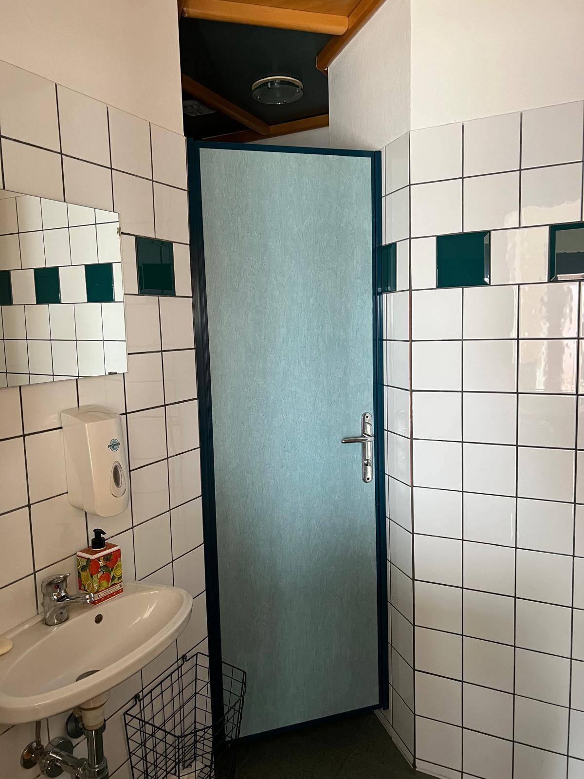 Weitere Ansicht der öffentlichen Toilette mit weißen Fliesen, Waschbecken und einer Kabinentür.