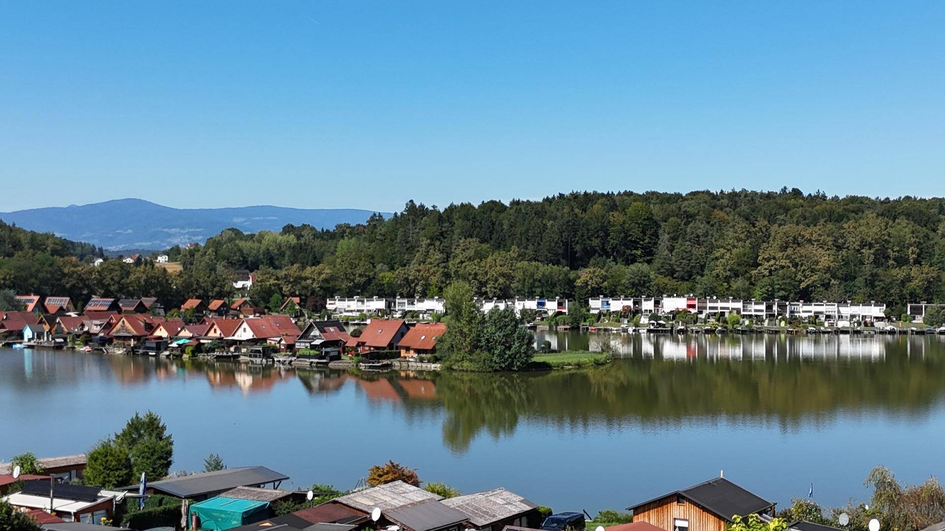 Panoramablick über den See mit Häusern am Ufer und bewaldeten Hügeln im Hintergrund.