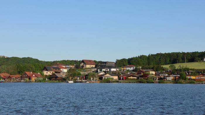 Panoramablick auf die Häuserreihe entlang des Seeufers mit bewaldetem Hintergrund.