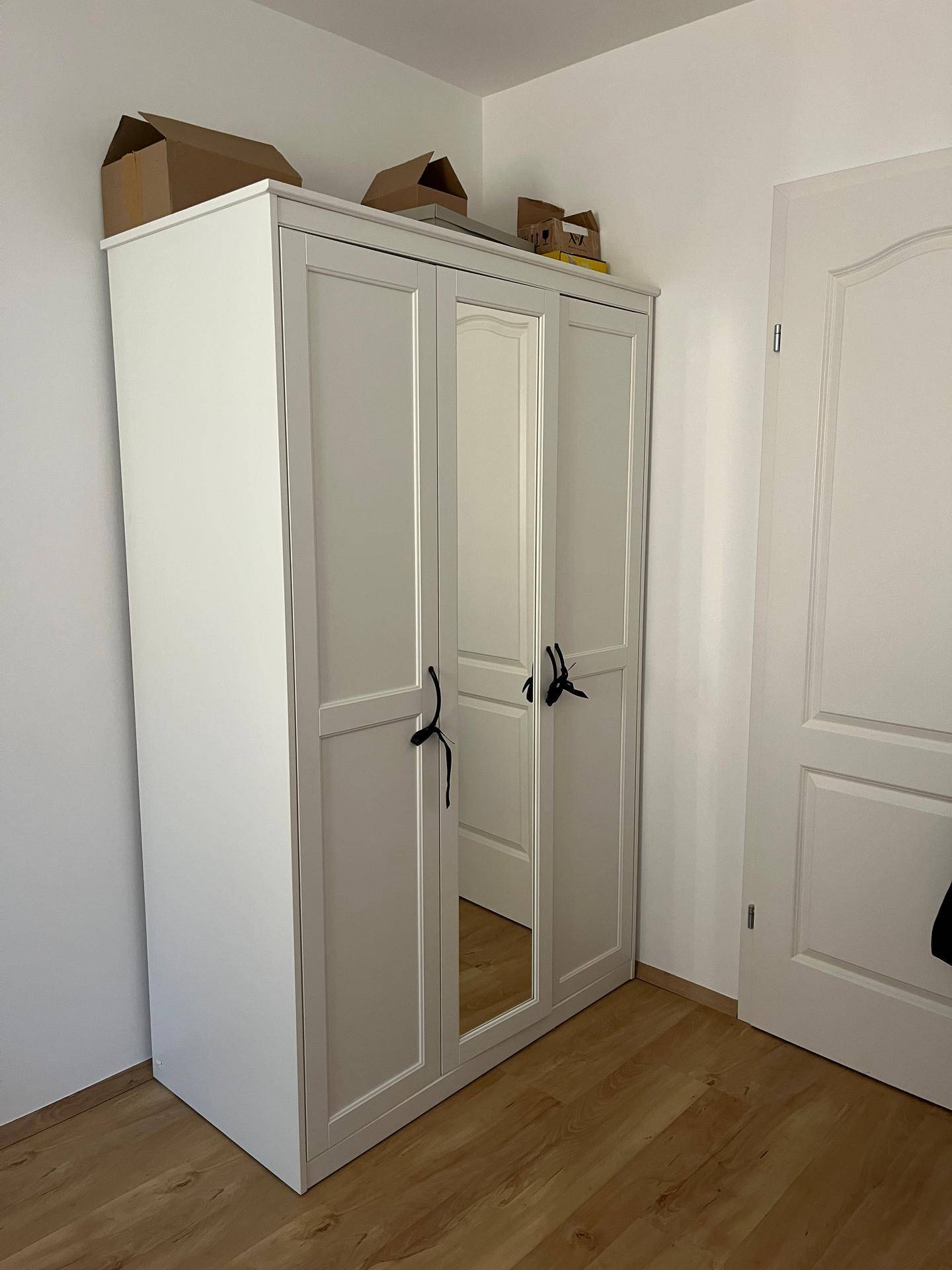 Weißer Kleiderschrank mit Spiegeltür in einem hellen Raum mit Holzboden.
