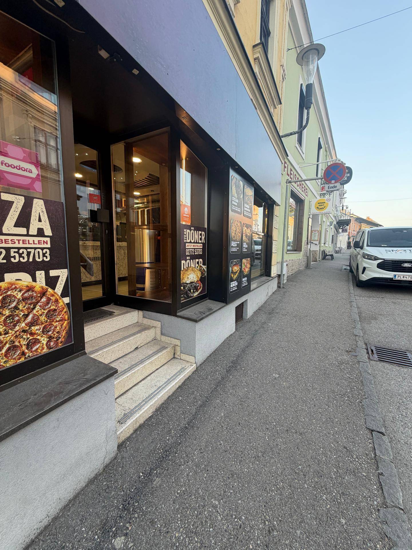 Blick auf die Ladenfront eines Restaurants mit Werbung für Pizza und Döner, sowie einem geparkten Lieferwagen.
