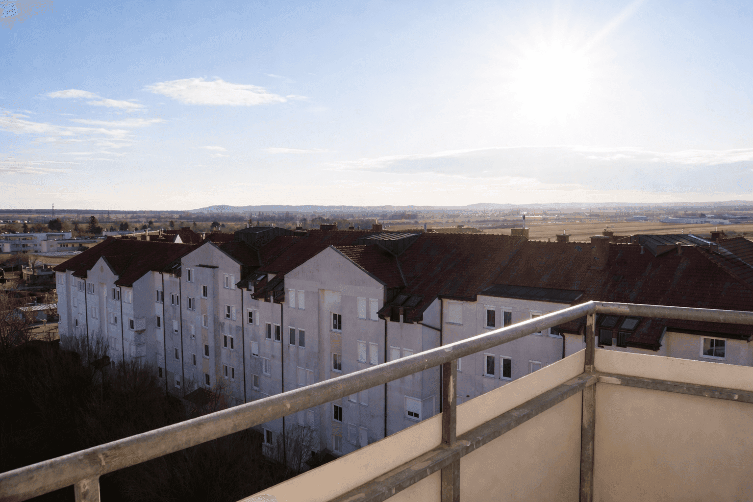 Weitblick vom Balkon über die umliegenden Wohngebäude und die weite Landschaft unter blauem Himmel.