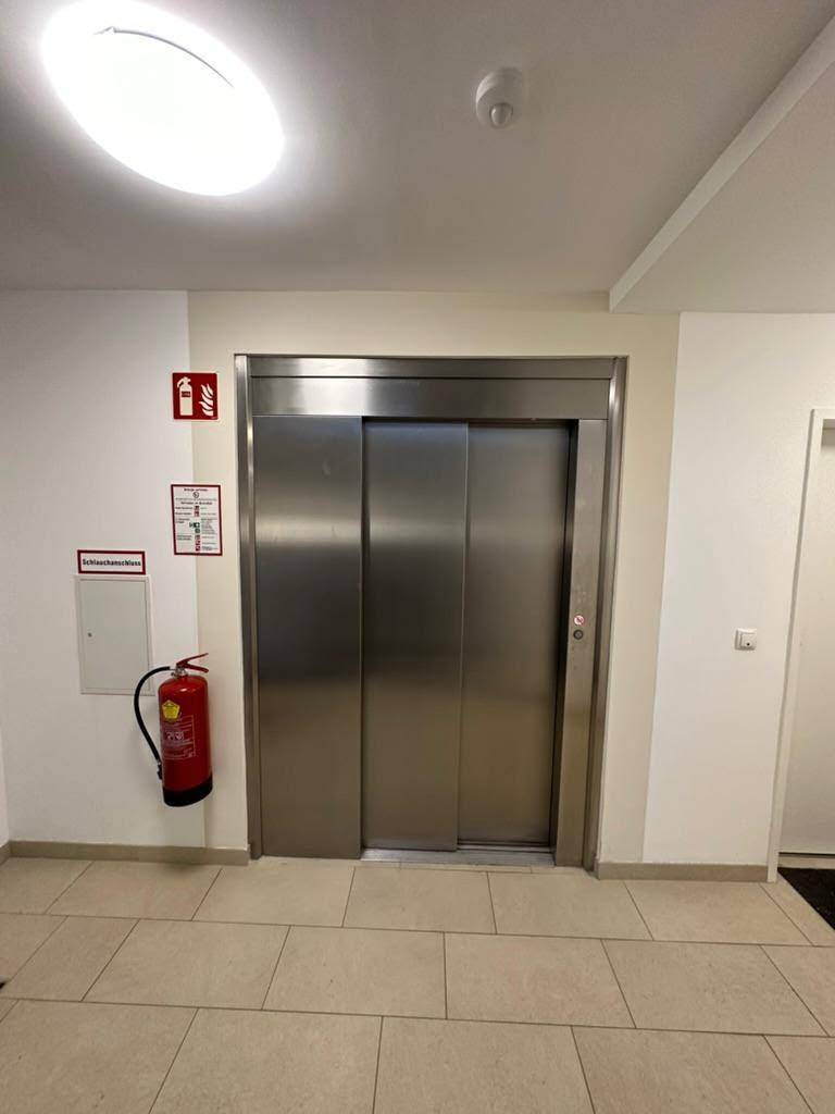Moderner Aufzug im Eingangsbereich des Gebäudes mit Fliesenboden und Brandschutzhinweisen.