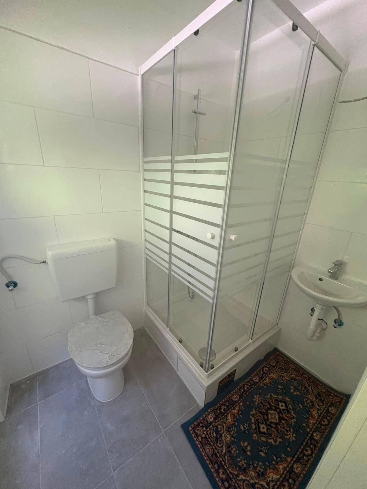 Kleines Badezimmer mit Duschkabine, Toilette, Waschbecken und hellgrauen Fliesen.