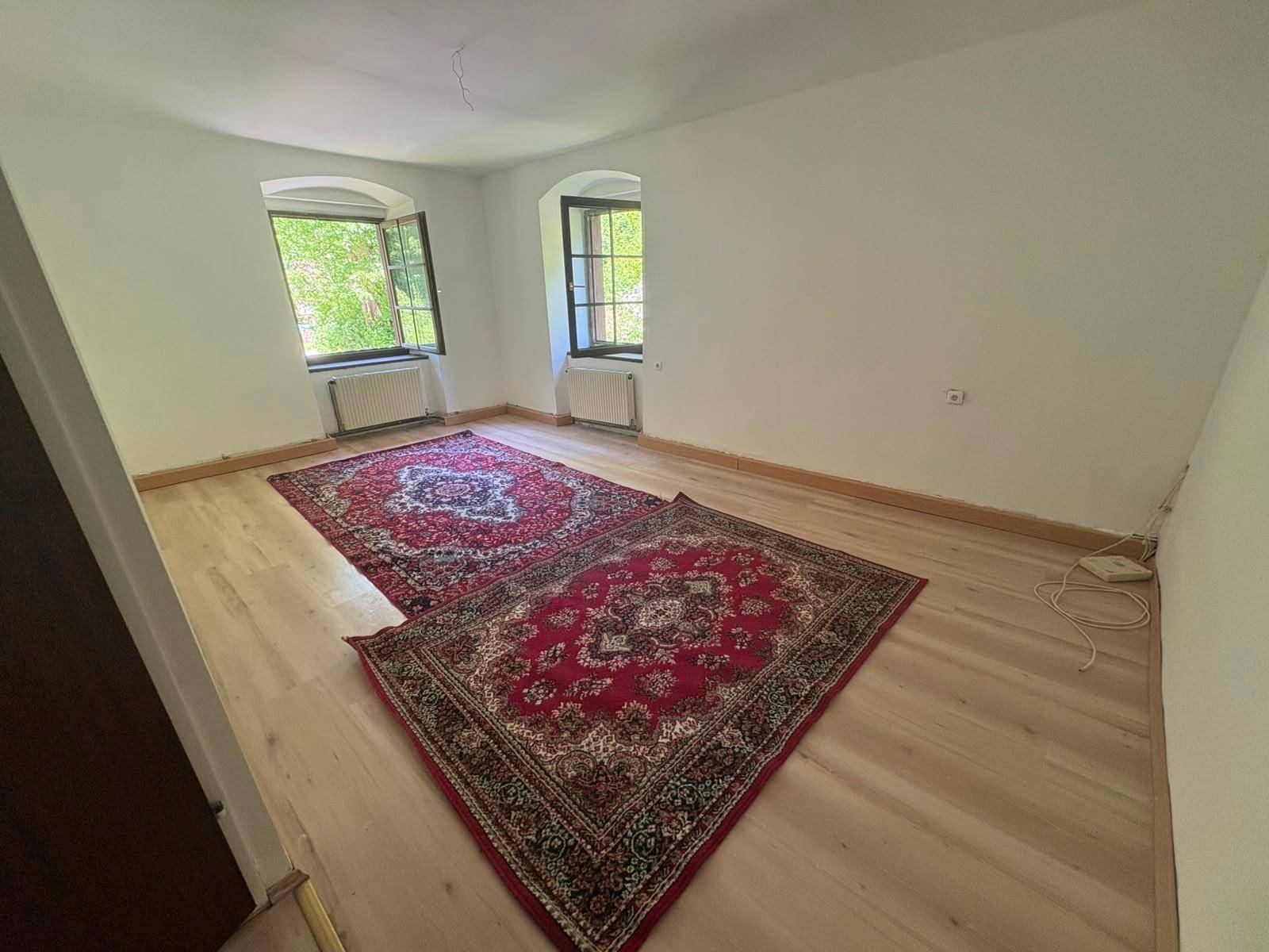 Helles Zimmer mit zwei Fenstern, Holzboden und zwei traditionellen roten Teppichen.