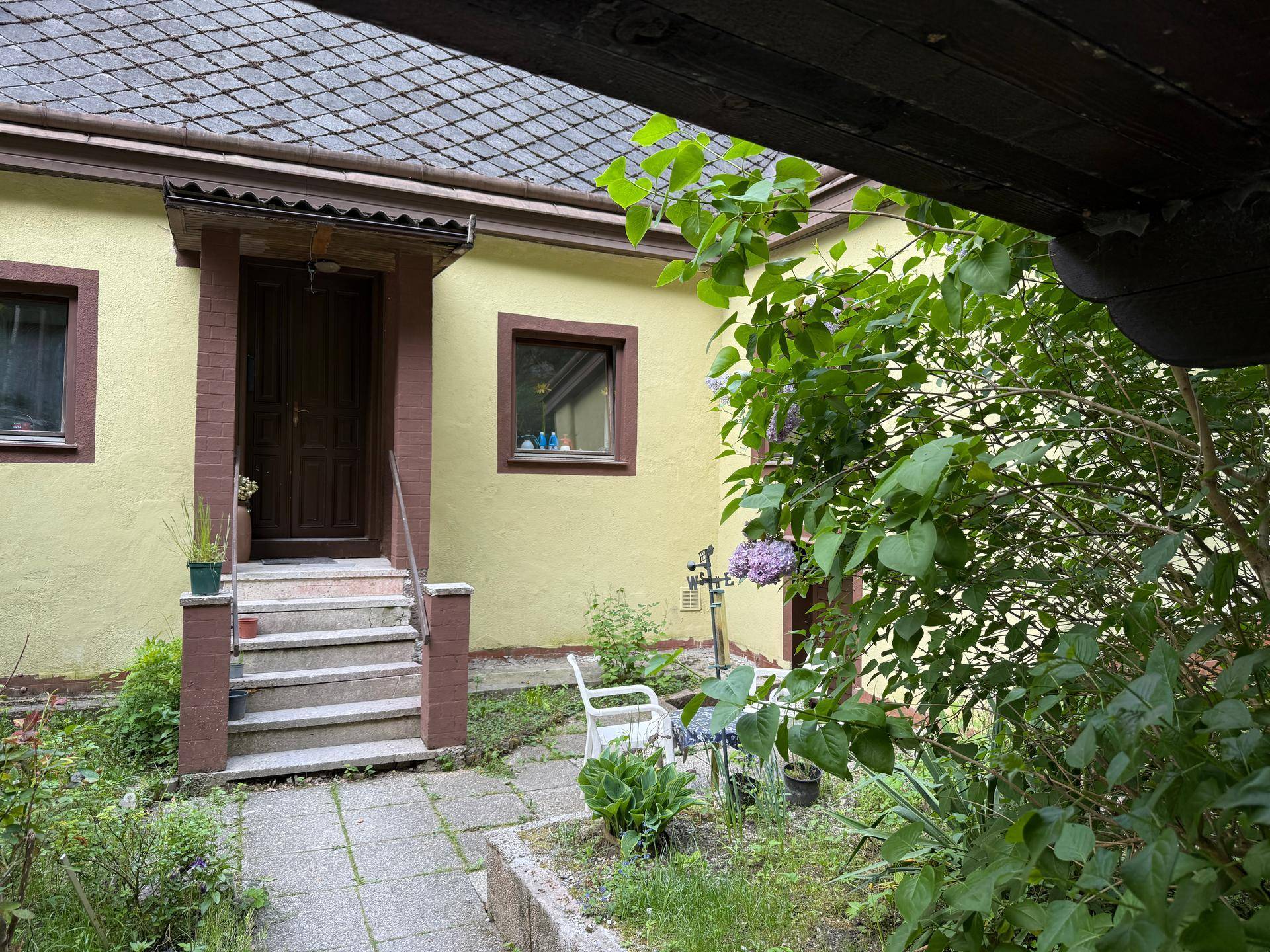 Eingangsbereich des Hauses mit Steinstufen, überdachtem Vordach und einer kleinen Terrasse mit Sitzgelegenheiten.
