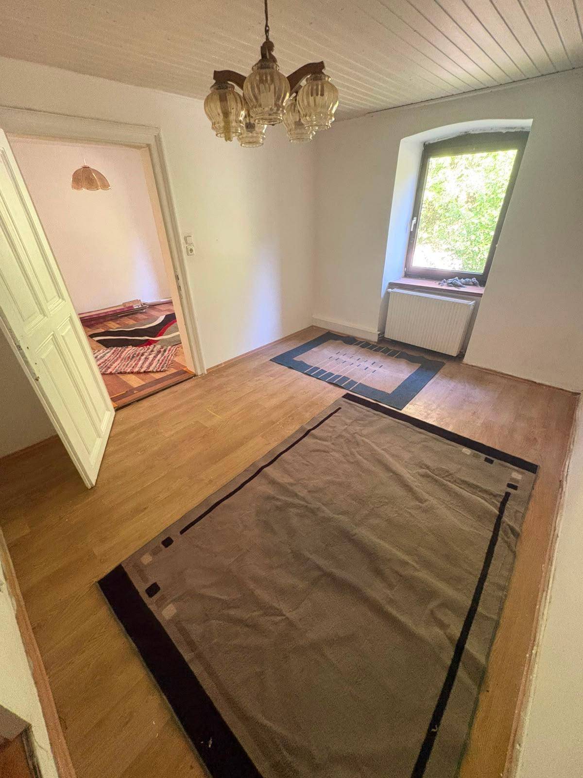 Helles Zimmer mit Holzboden und zwei Teppichen, das durch eine offene Tür mit einem weiteren Raum verbunden ist.
