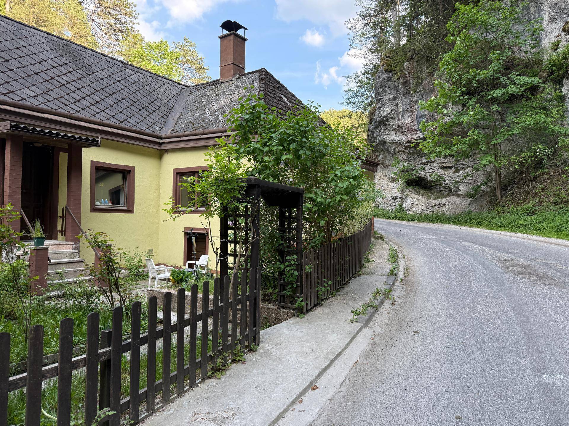 Seitenansicht des Hauses mit Holzzaun und einer kurvigen Landstraße, umgeben von Felsen und Bäumen.