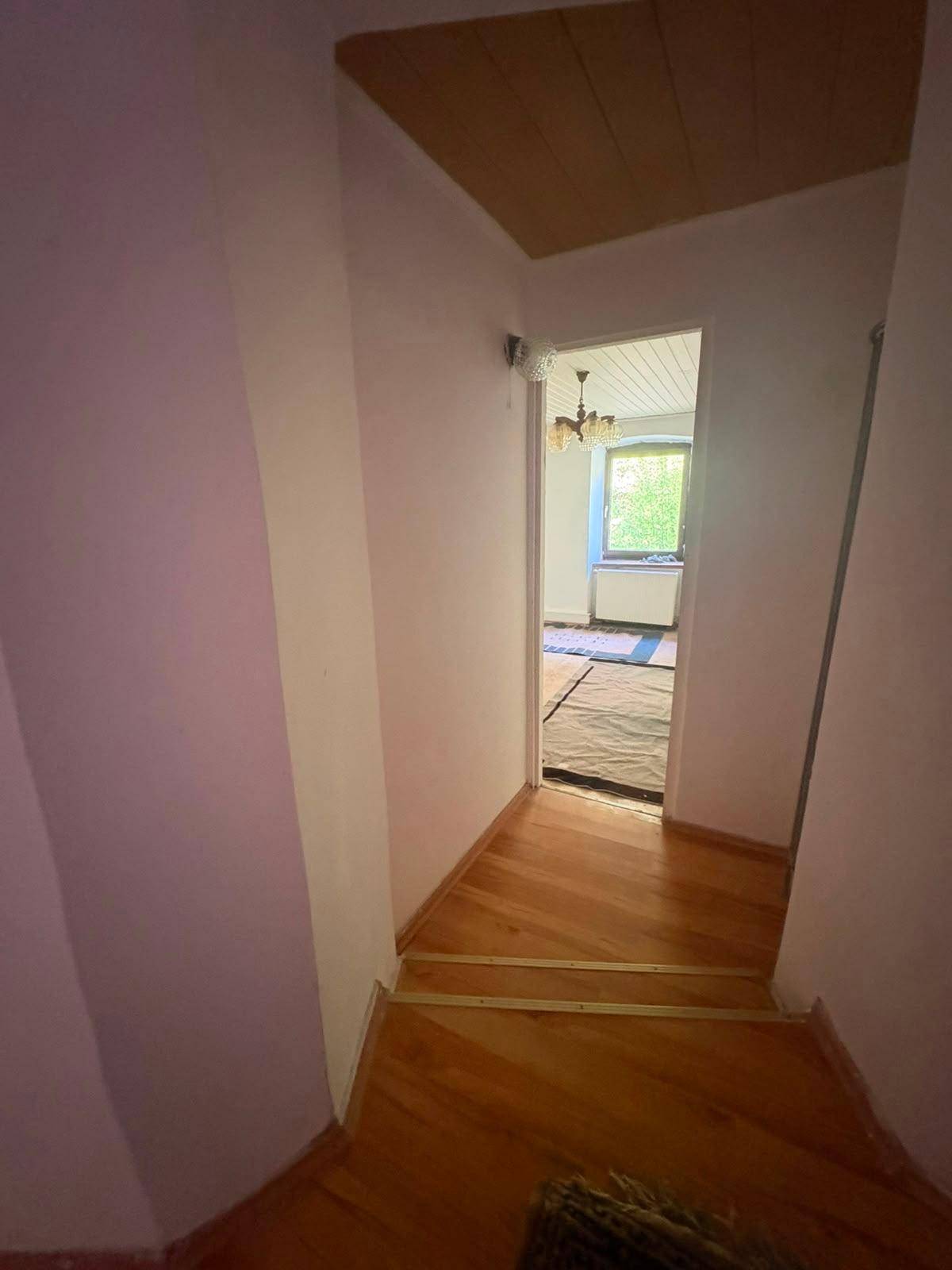 Schmaler Flur mit Holzboden, der zu einem hellen Zimmer mit Fenster und Kronleuchter führt.
