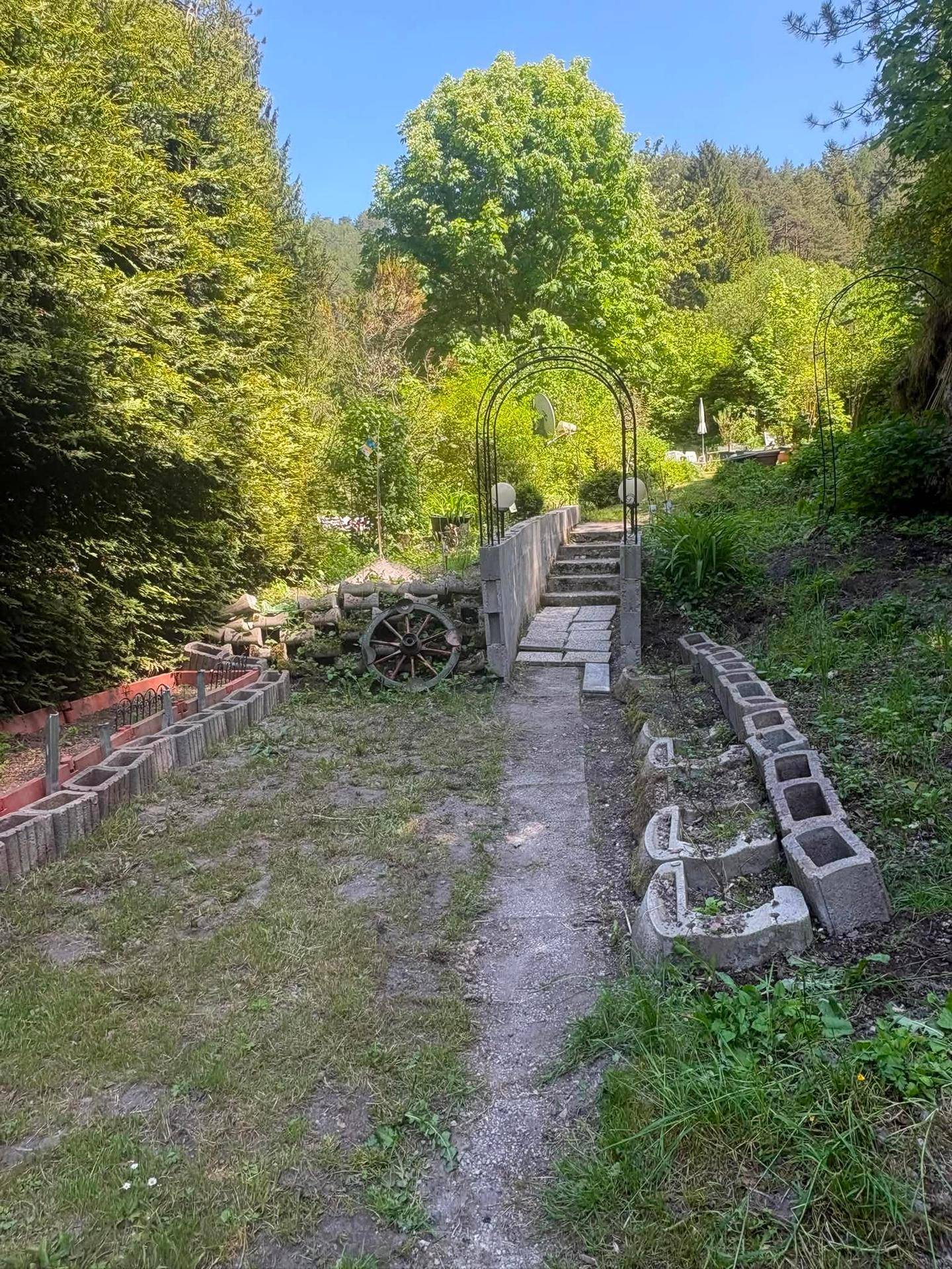 Gartenweg mit Betonstufen, umgeben von Bäumen und dekorativen Elementen wie einem alten Wagenrad.