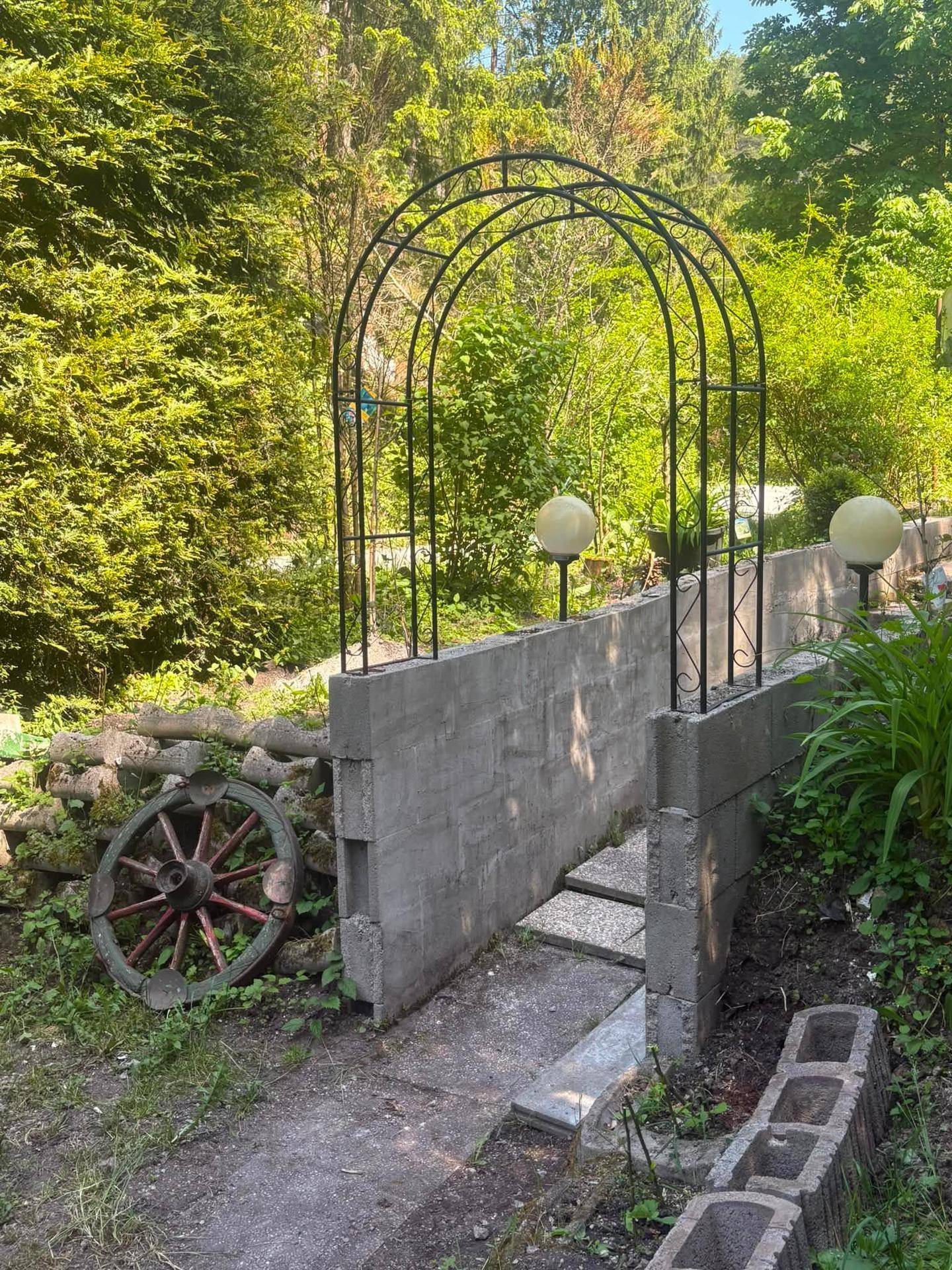 Metallener Gartenbogen über Steinstufen, flankiert von Gartenleuchten und üppigem Grün.