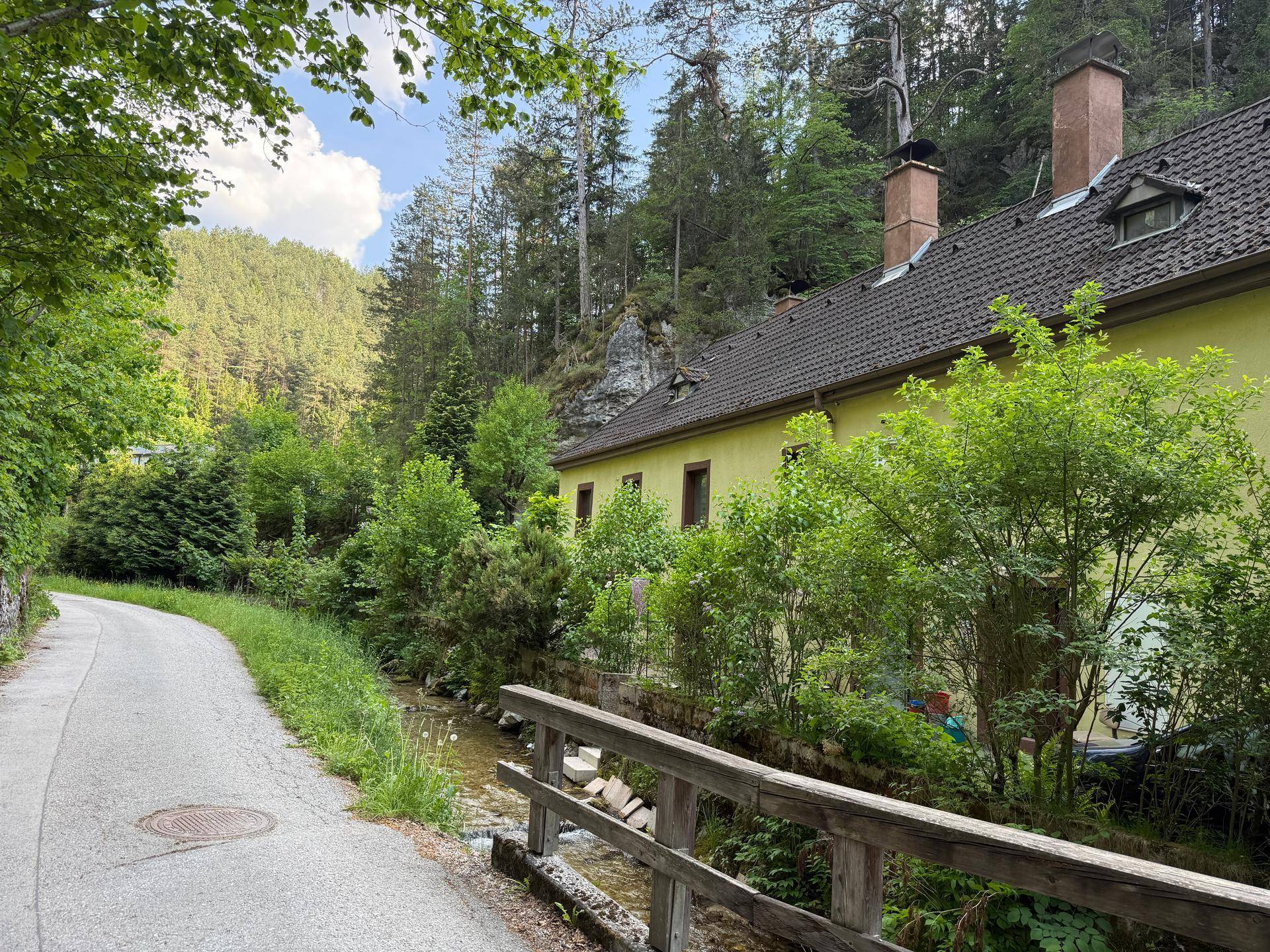 Hausansicht von der Straße aus, mit einem Bach im Vordergrund und bewaldeten Hügeln im Hintergrund.