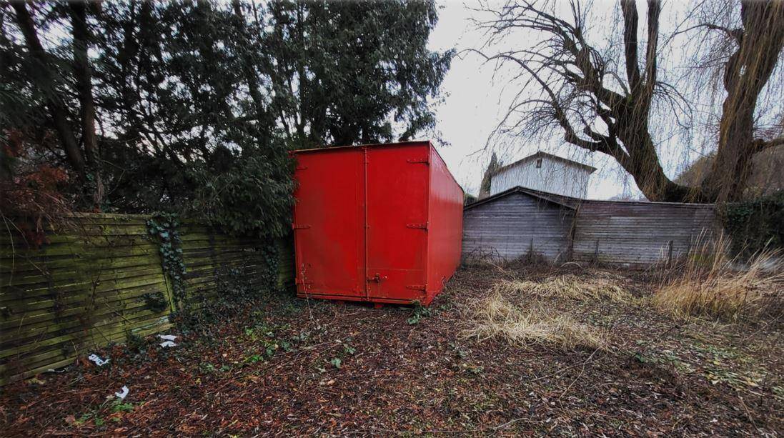 Roter Metallcontainer steht auf einer grasbewachsenen Fläche neben einem Holzzaun und Bäumen.