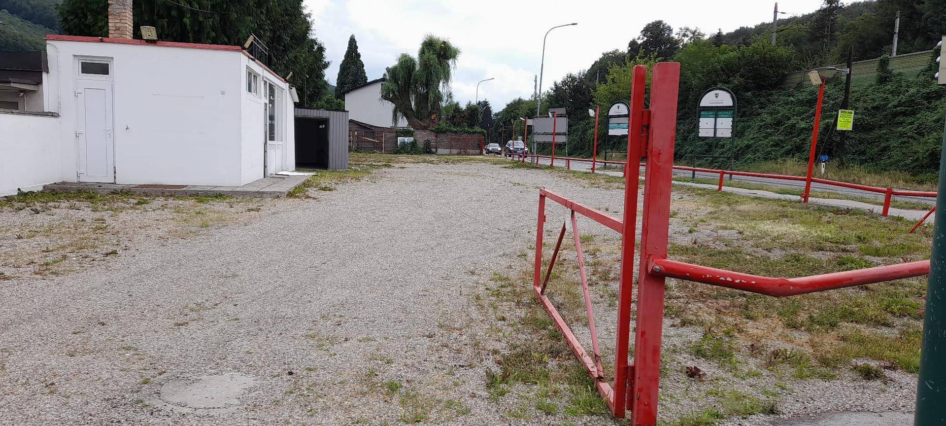 Weitläufiger Schotterparkplatz mit einem roten Tor, einem kleinen Gebäude und einer Straße im Hintergrund.