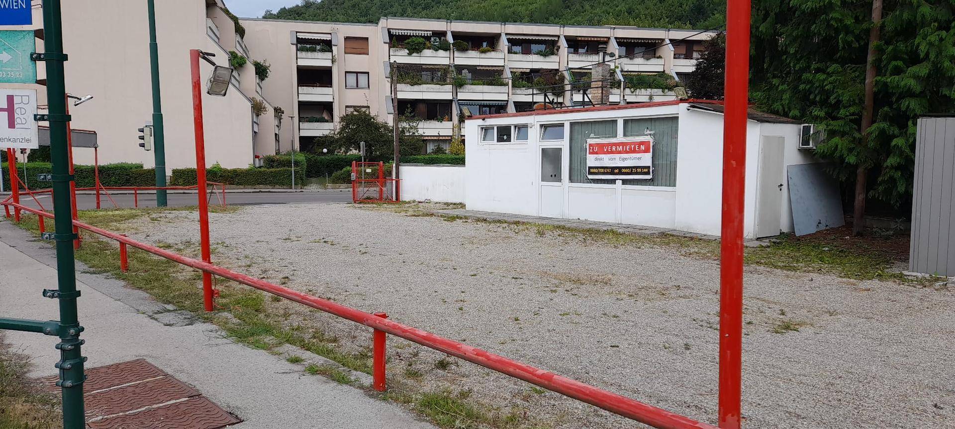 Schotterfläche mit einem kleinen weißen Gebäude, das ein "ZU VERMIETEN"-Schild trägt, und einem Mehrfamilienhaus im Hintergrund.