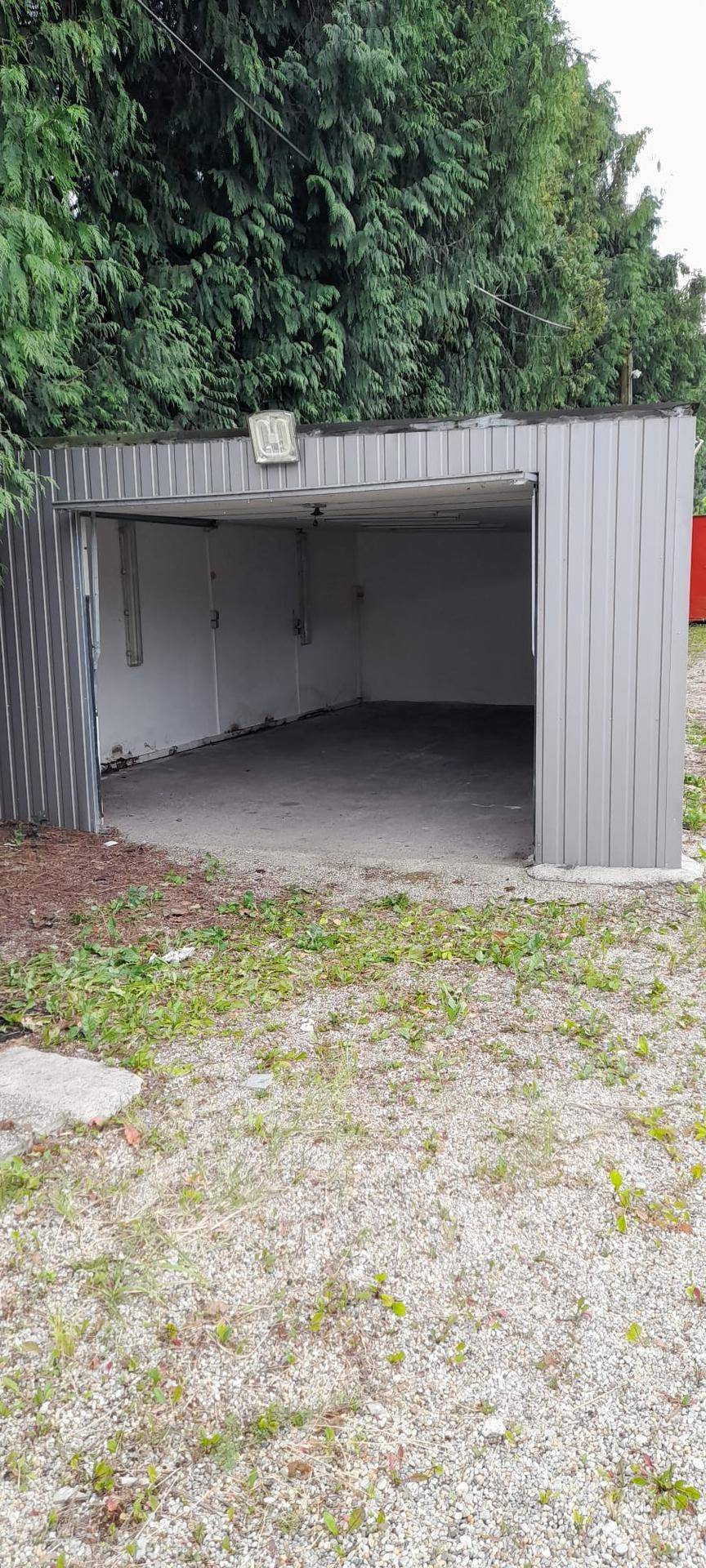 Offene graue Metallgarage mit Betonboden, umgeben von Bäumen und einer Schotterfläche.