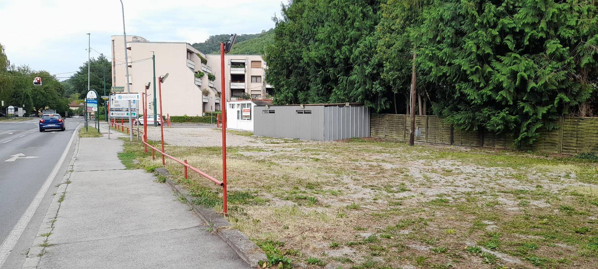 Große Schotterfläche mit einem grauen Metallschuppen, umgeben von Bäumen und einer Straße.