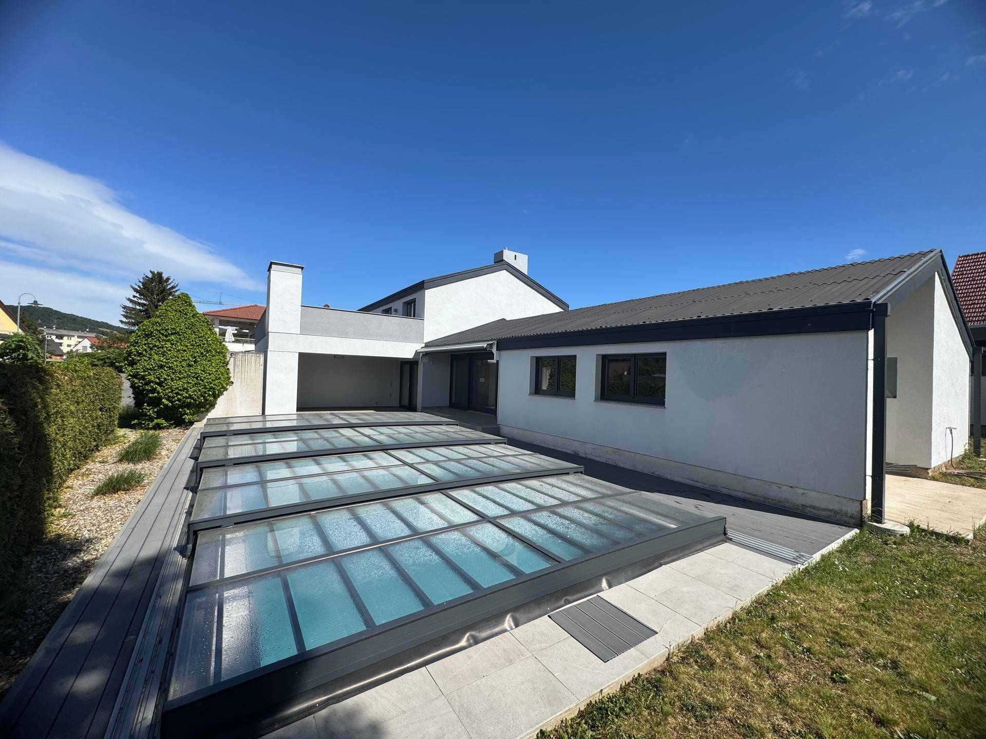 Ansicht des modernen Hauses mit abgedecktem Pool und angrenzender Rasenfläche im Sonnenschein.