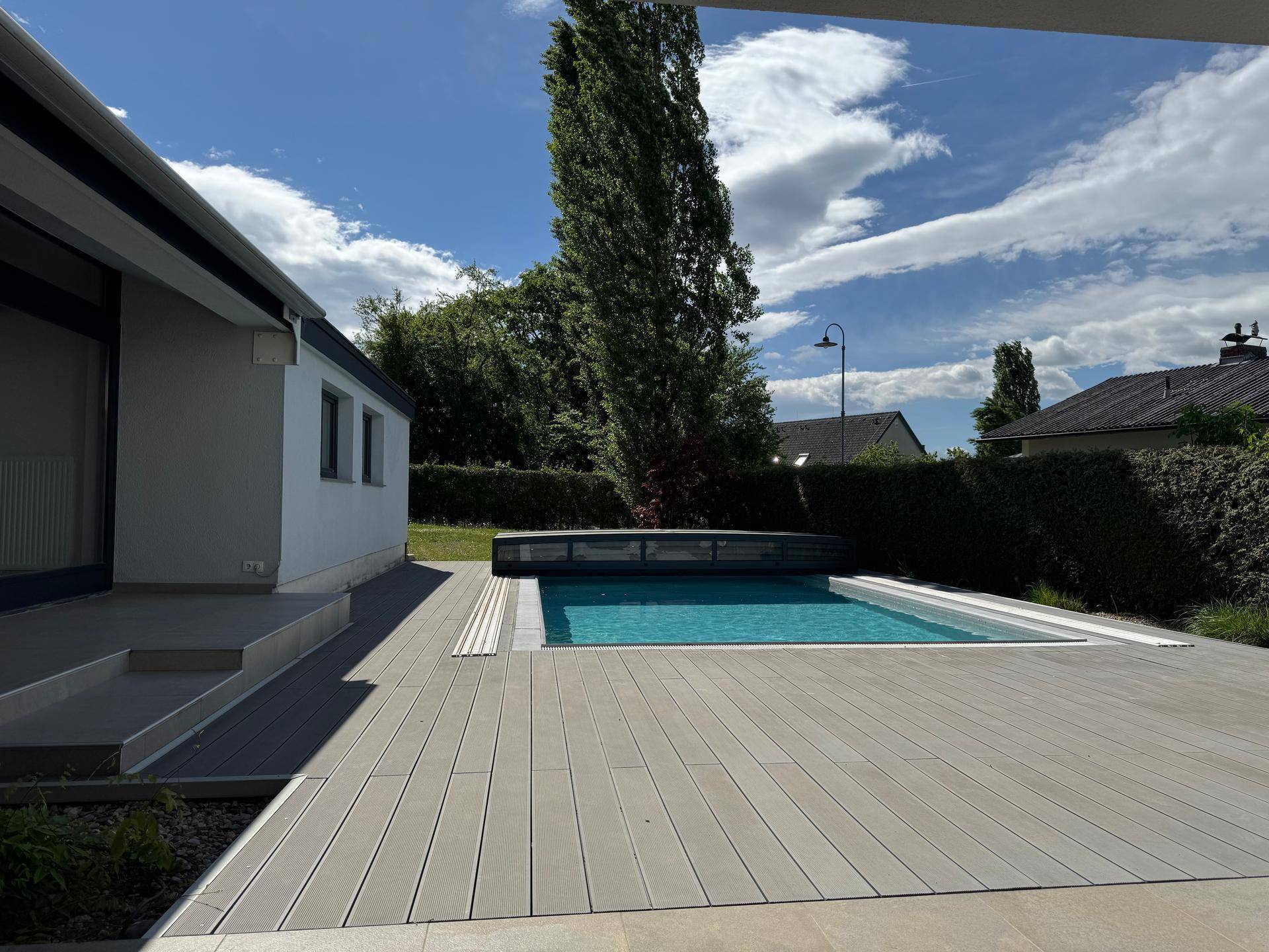 Moderner Außenbereich mit Swimmingpool, Holzterrasse und gepflegtem Garten unter blauem Himmel.