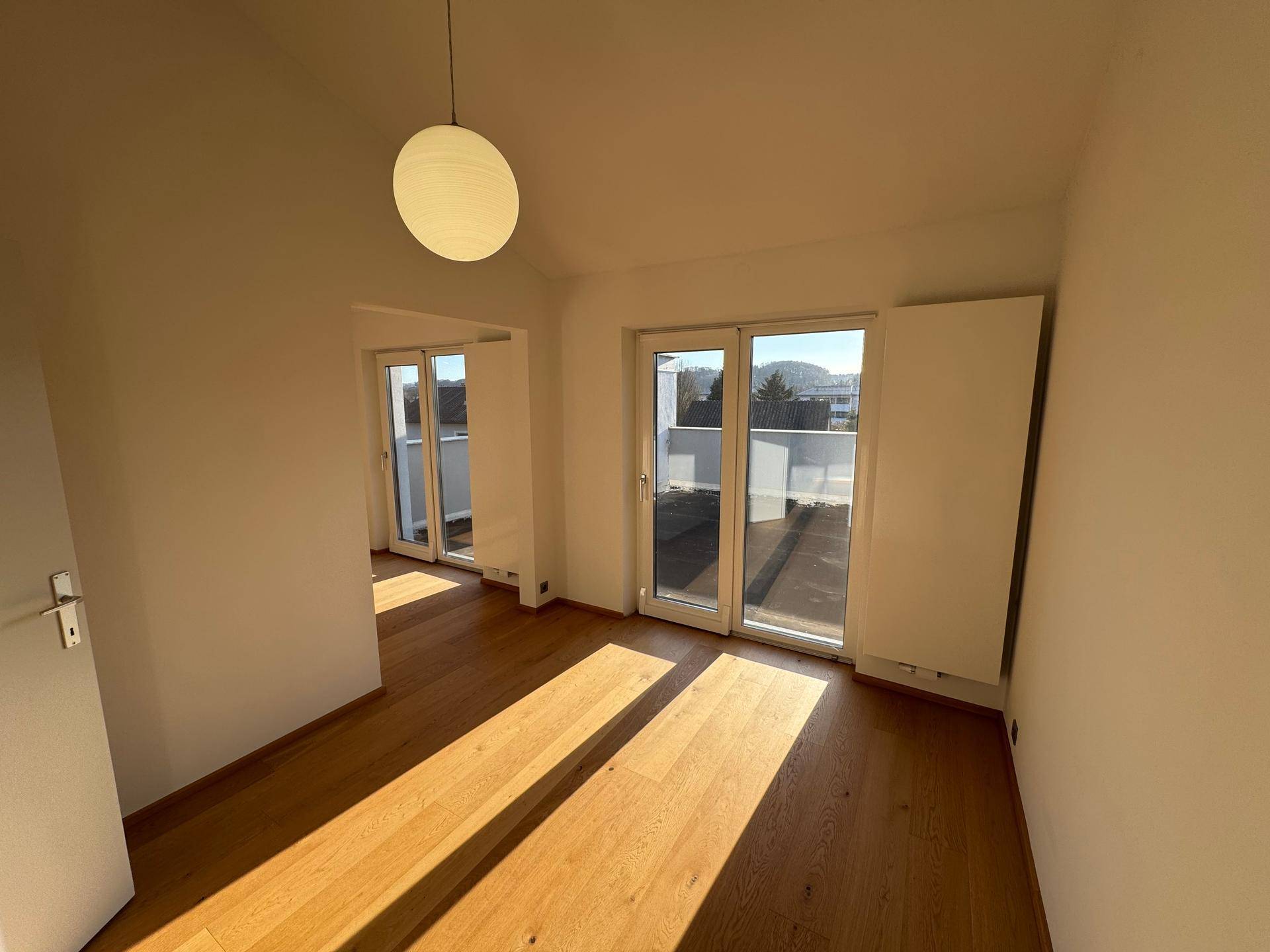 Sonniger Raum mit Holzboden und zwei Balkontüren, die viel Licht hereinlassen.