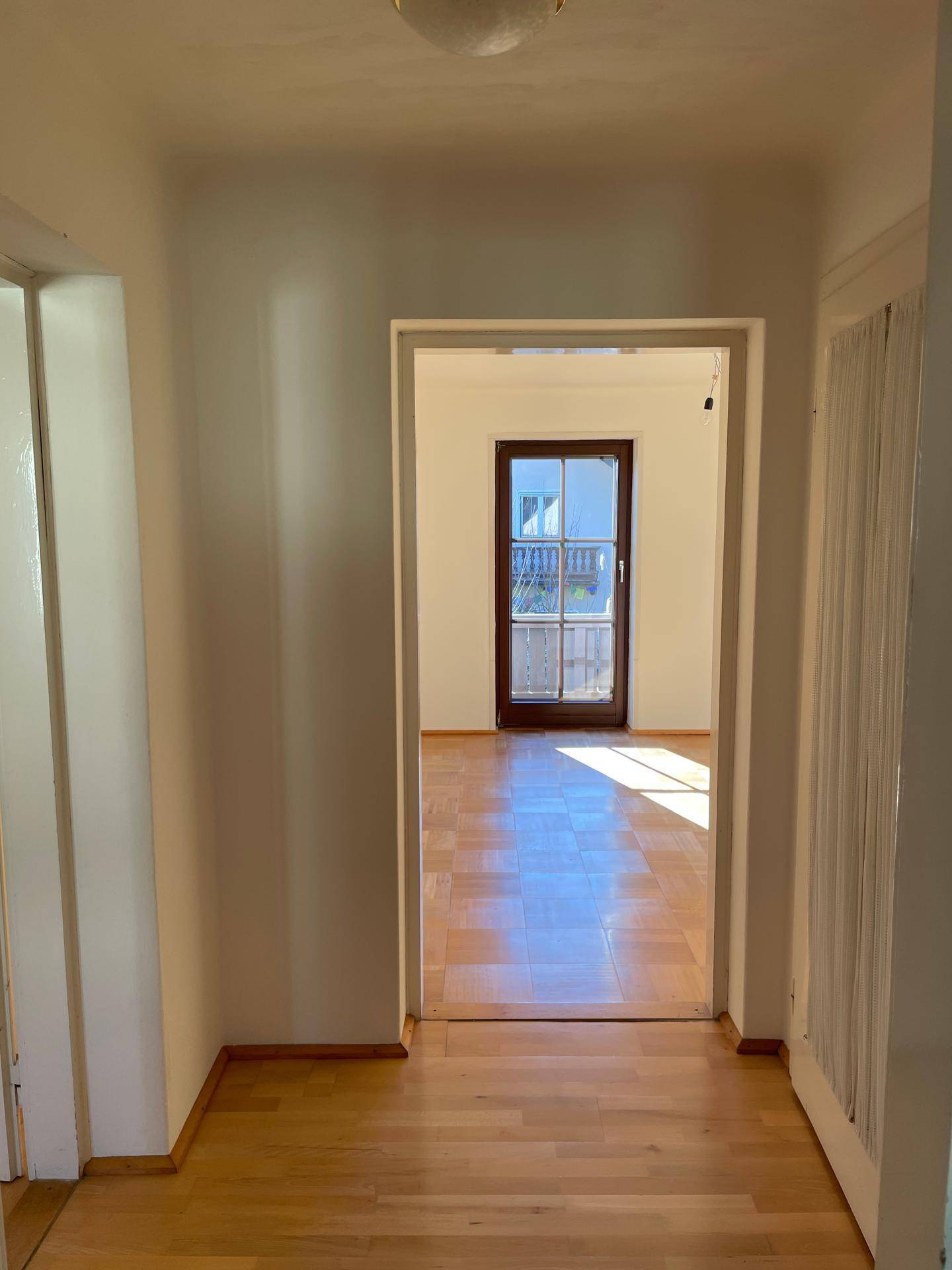 Heller Flur mit Parkettboden und Durchgang zu einem Zimmer mit Balkonzugang.