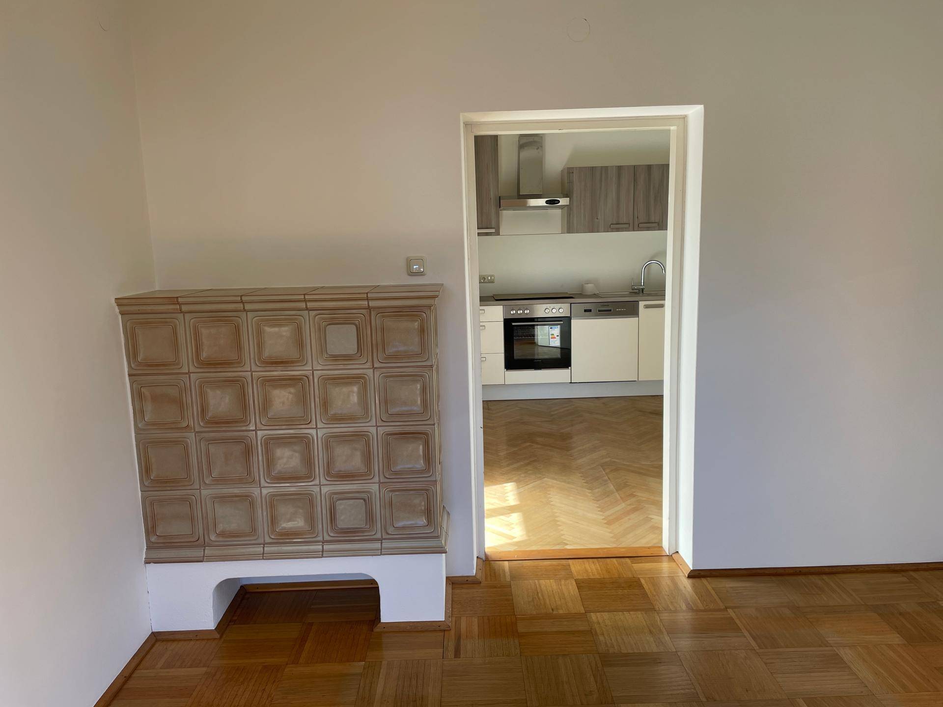Wohnzimmer mit Kachelofen und Durchgang zur Küche, ausgestattet mit Parkettboden.