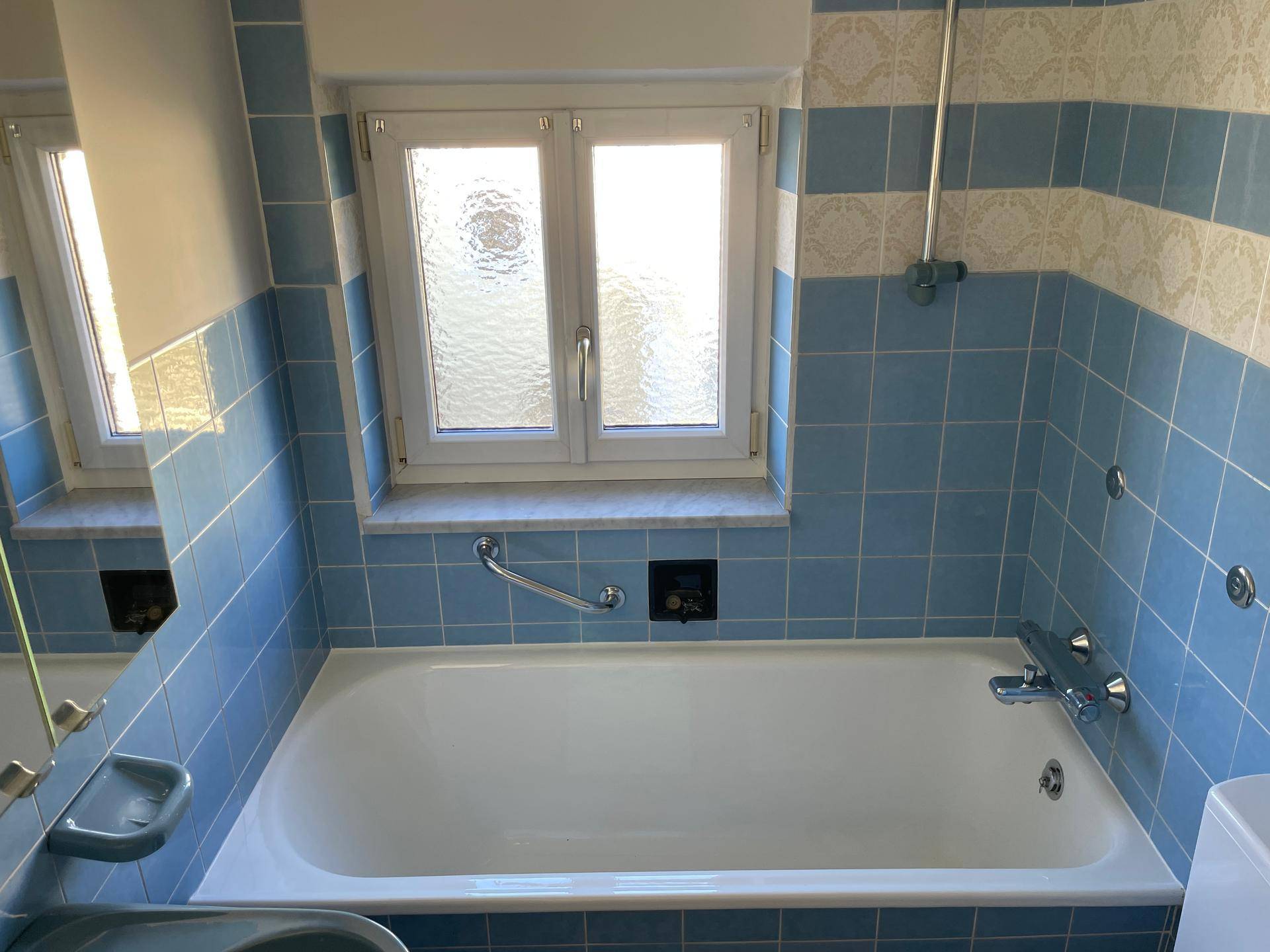 Badezimmer mit Badewanne, blauen Wandfliesen und Fenster für Tageslicht.