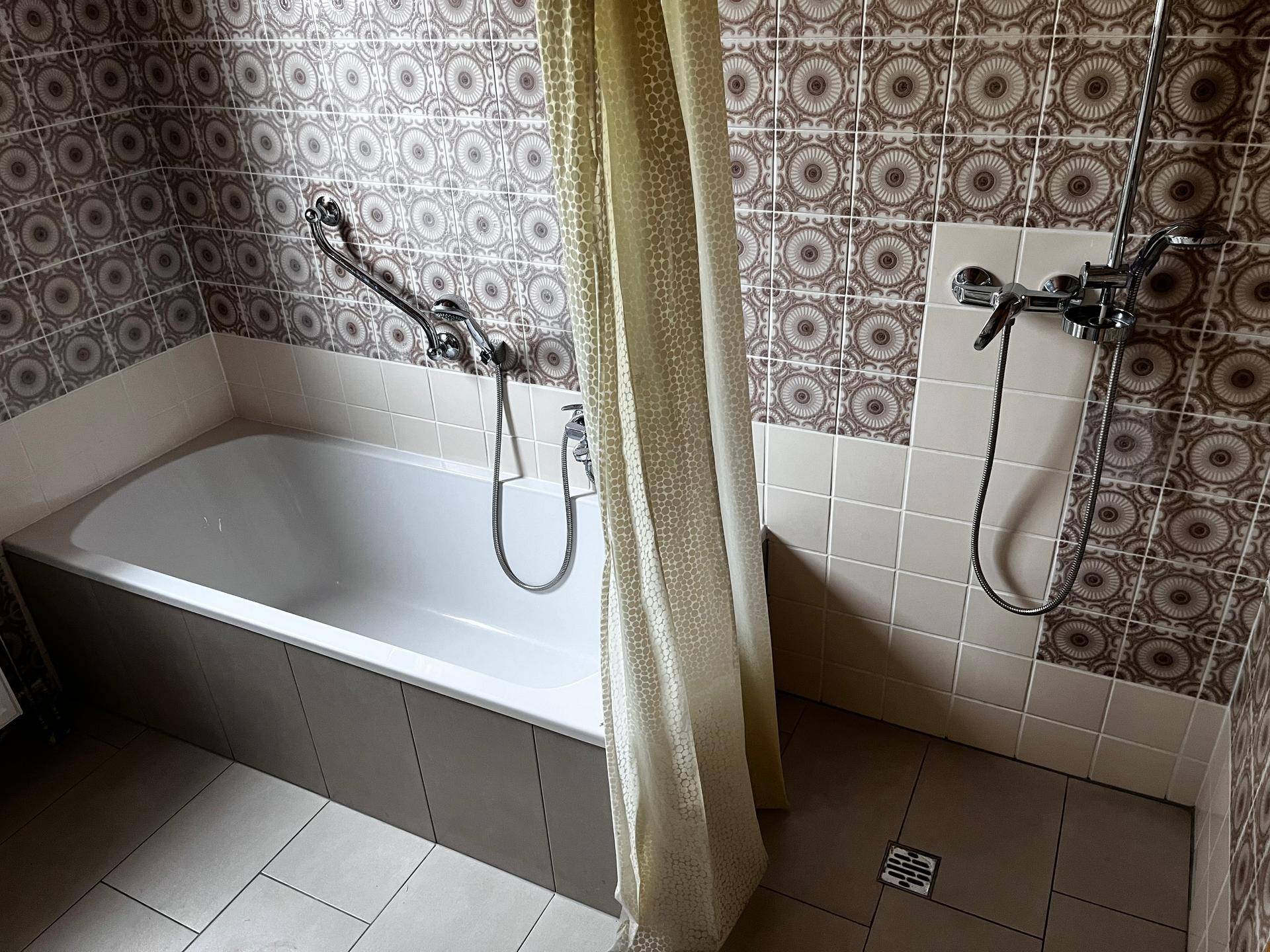 Badezimmer mit Badewanne, Duschvorrichtung und gemusterten Wandfliesen in Brauntönen.
