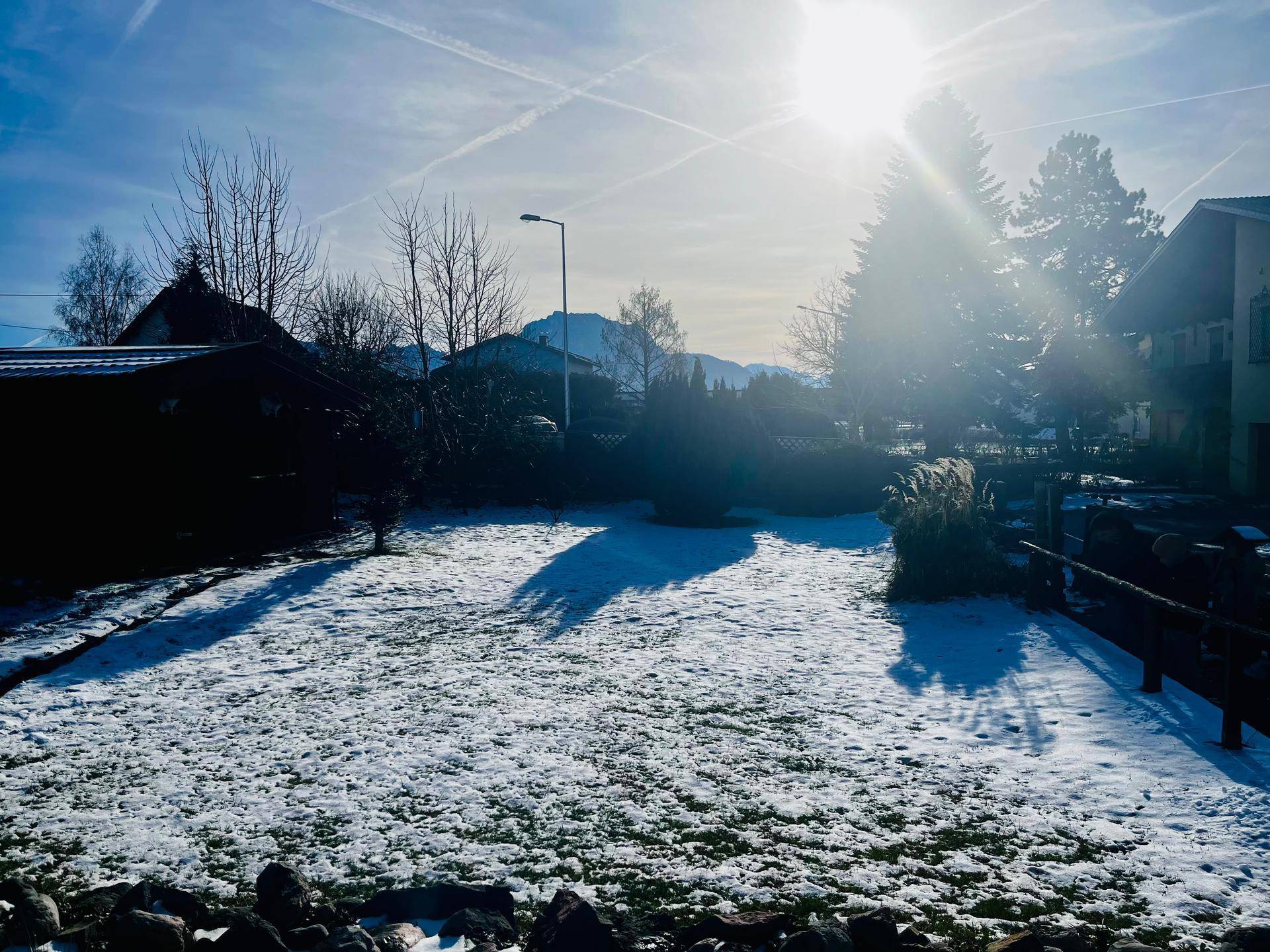 Verschneiter Gartenbereich mit Bäumen und Blick auf die Berge an einem sonnigen Wintertag.